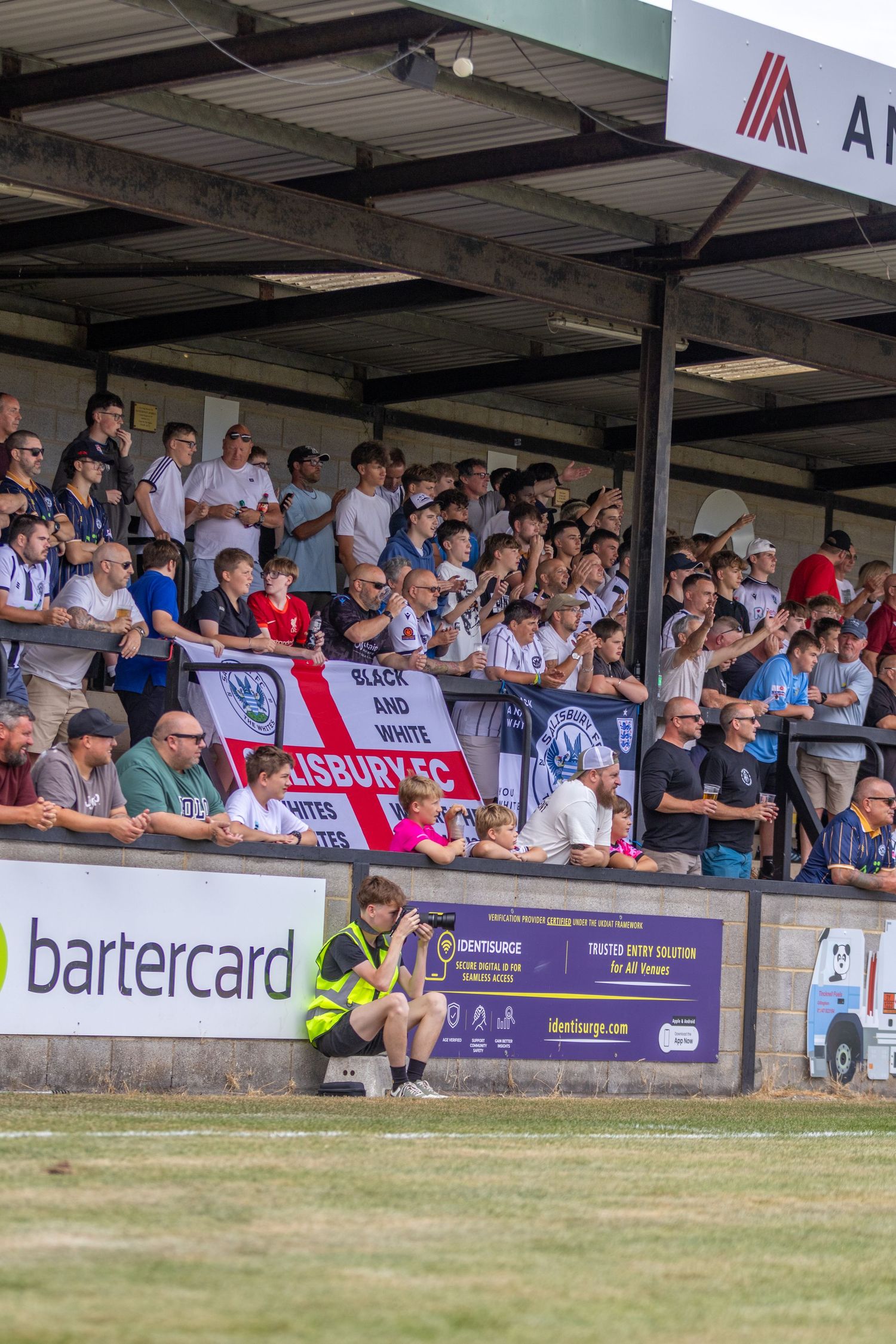 Salisbury FC fans fill a covered stand holding a black and white banner during a home non-league football match.