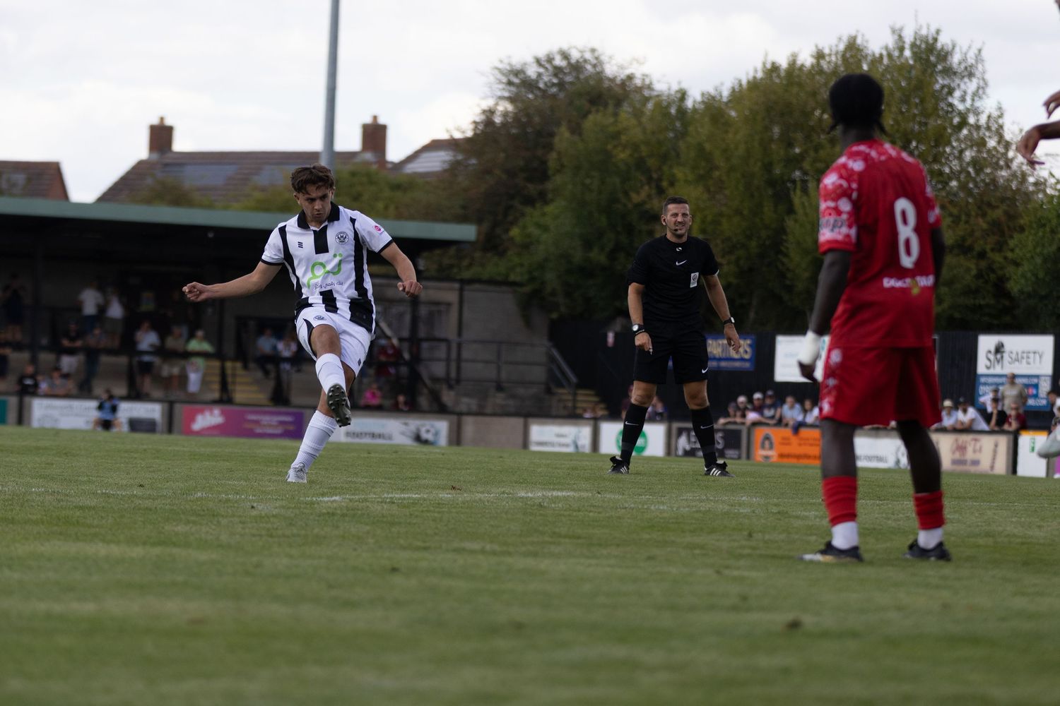 A footballer in black and white kit kicks the ball while a red-shirted opponent watches.