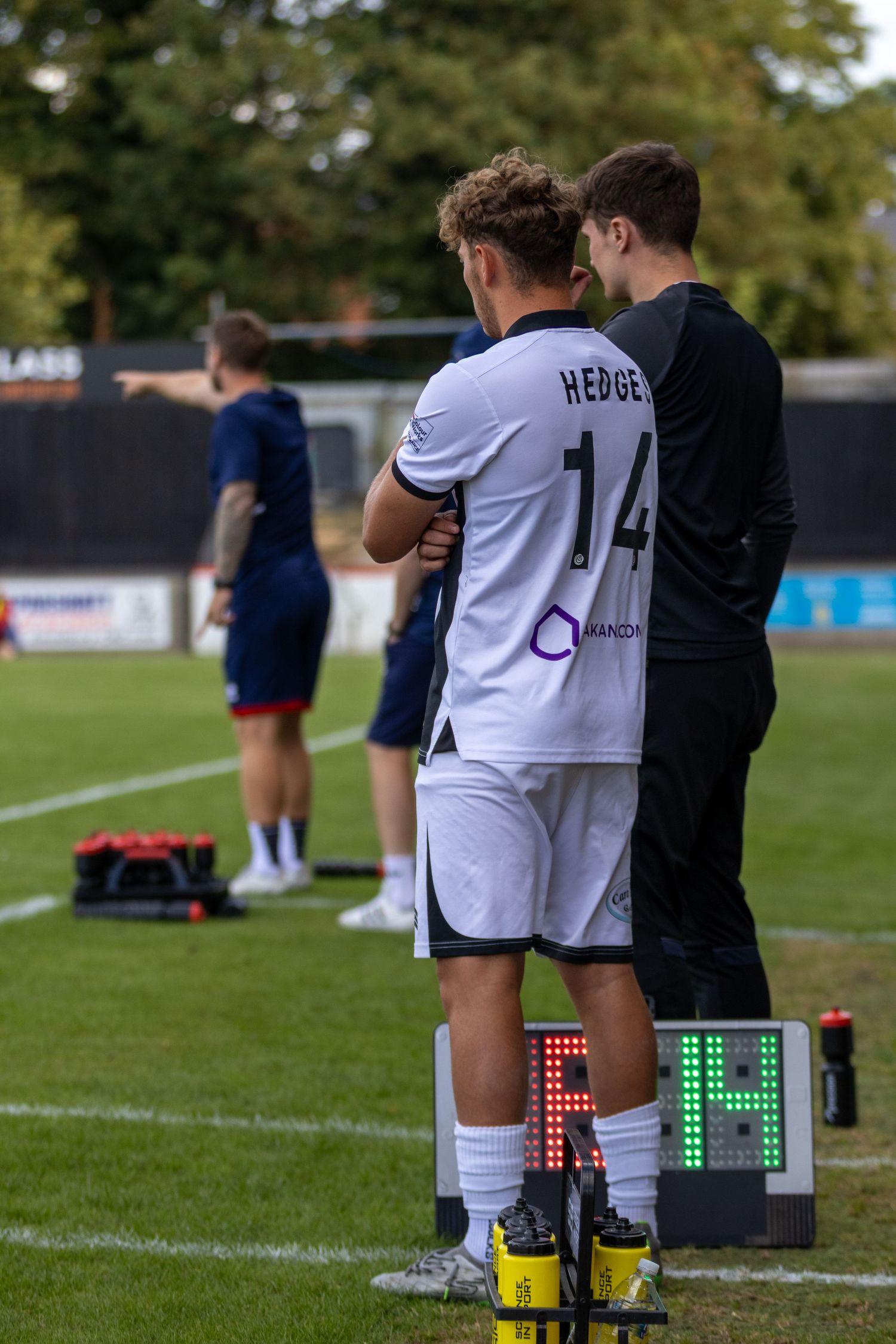 Soccer player number 14 Hedge stands on the sideline with a coach, ready to substitute during a football match.