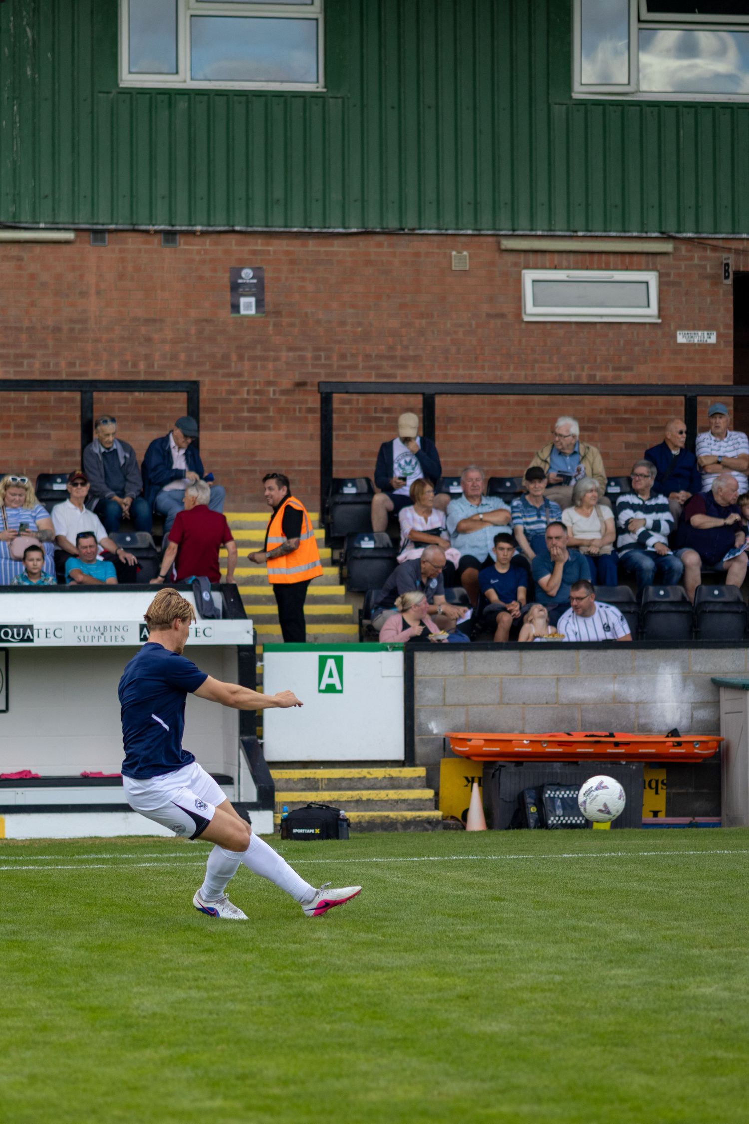 A footballer in navy blue kit strikes the ball powerfully during a match at a non-league football ground.