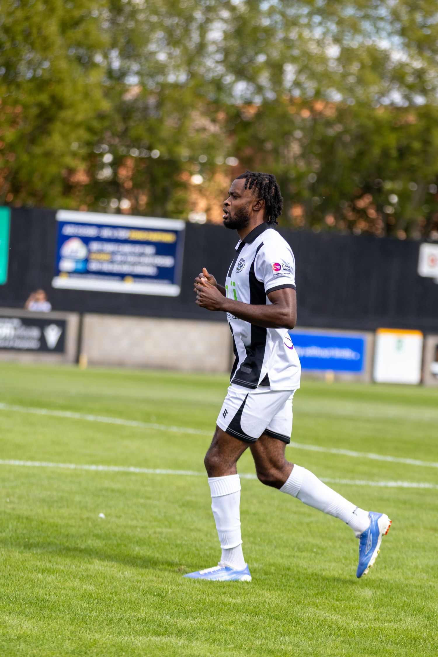 Football player in white and black kit with blue boots jogging across a green pitch.