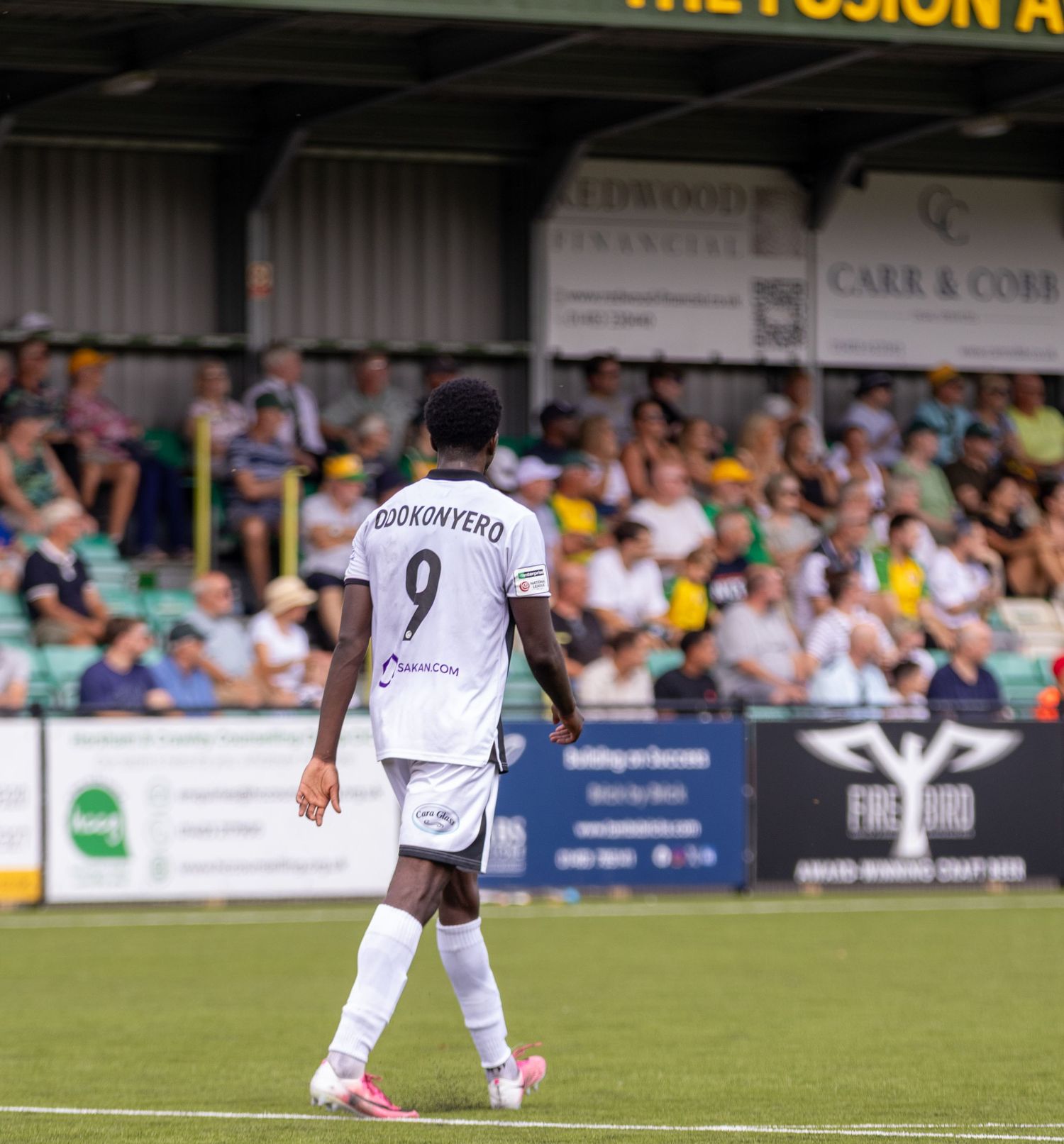 Footballer wearing number 9 Odukunyera white jersey walks on pitch before a crowded stadium stand.
