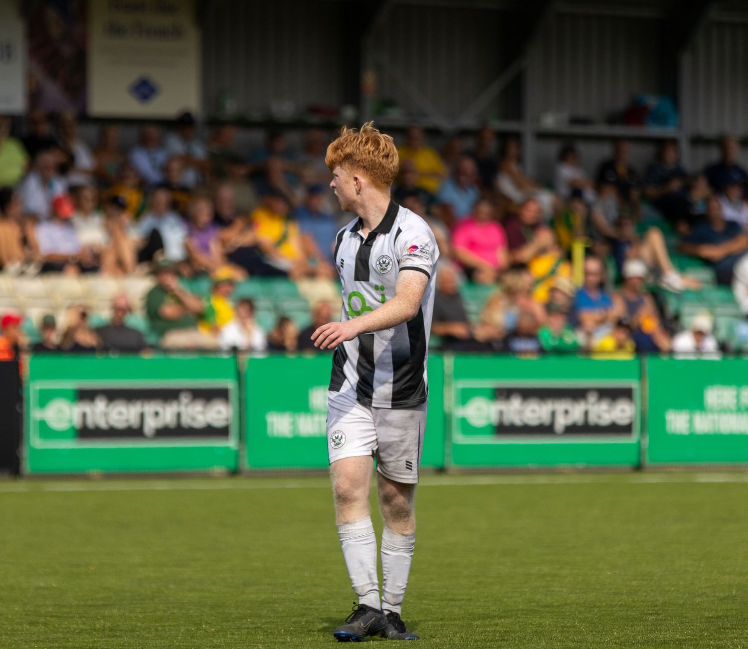 A young red-haired footballer in black and white striped kit stands on green pitch during a sunny match day.