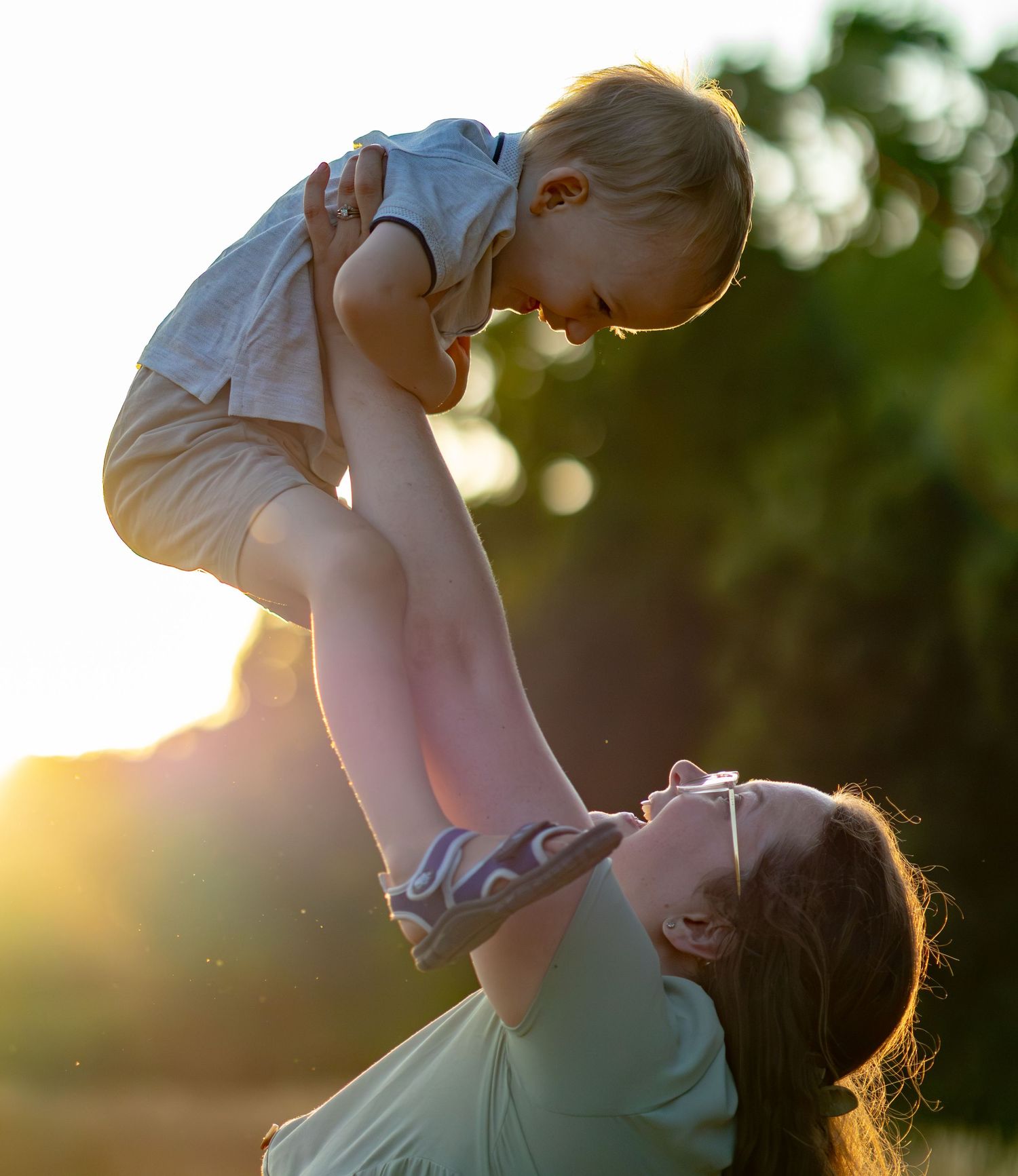 A joyful mother lifts her laughing toddler high in the air during a golden sunset outdoors.