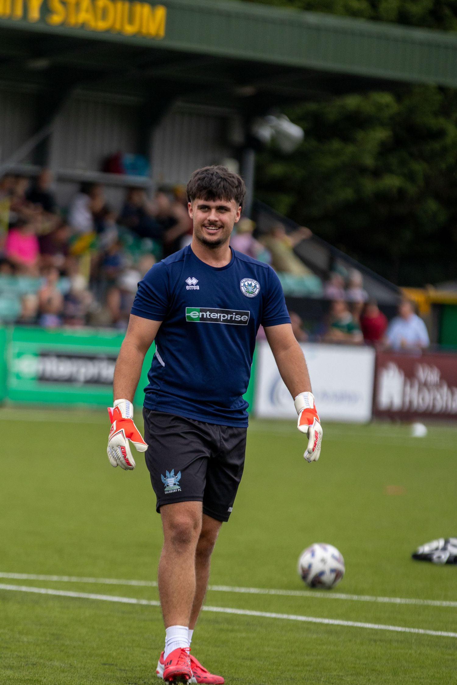 Smiling goalkeeper in navy Errea kit with Enterprise sponsor and white gloves during pre-match warmup at green stadium.