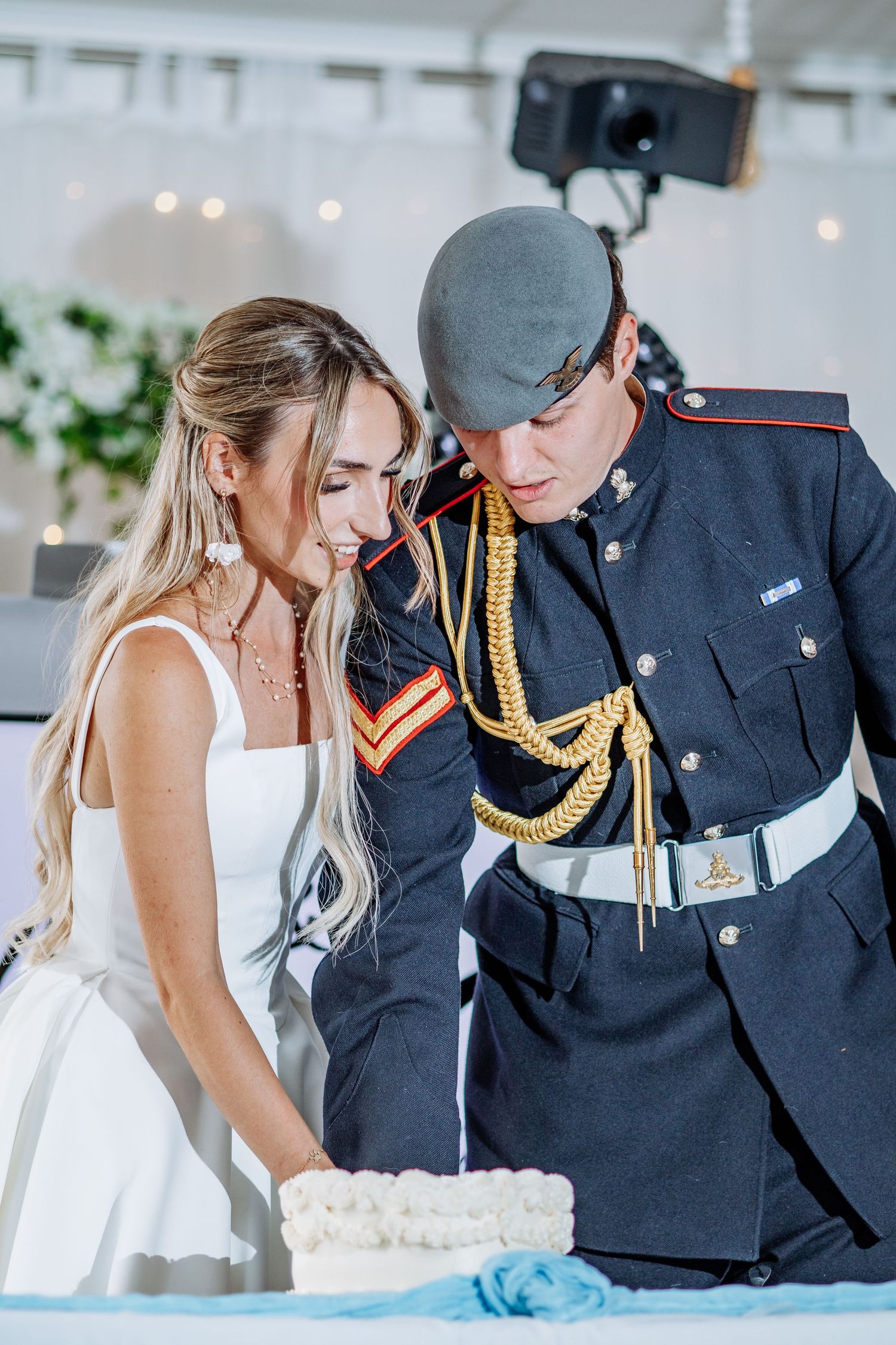 Bride in white dress and groom in military uniform cutting wedding cake together.