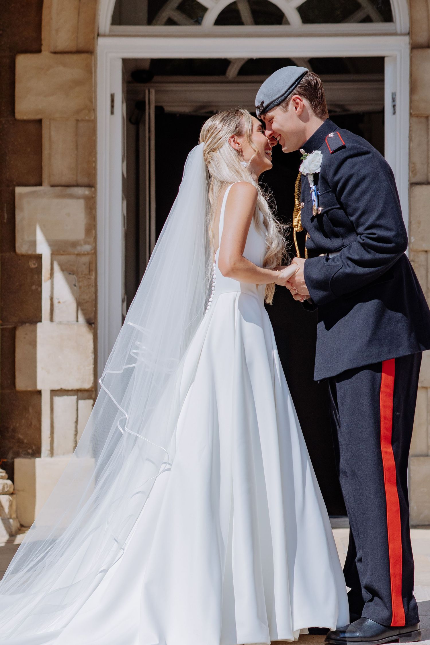Bride in elegant white gown and veil shares a kiss with groom in military dress uniform at venue entrance.