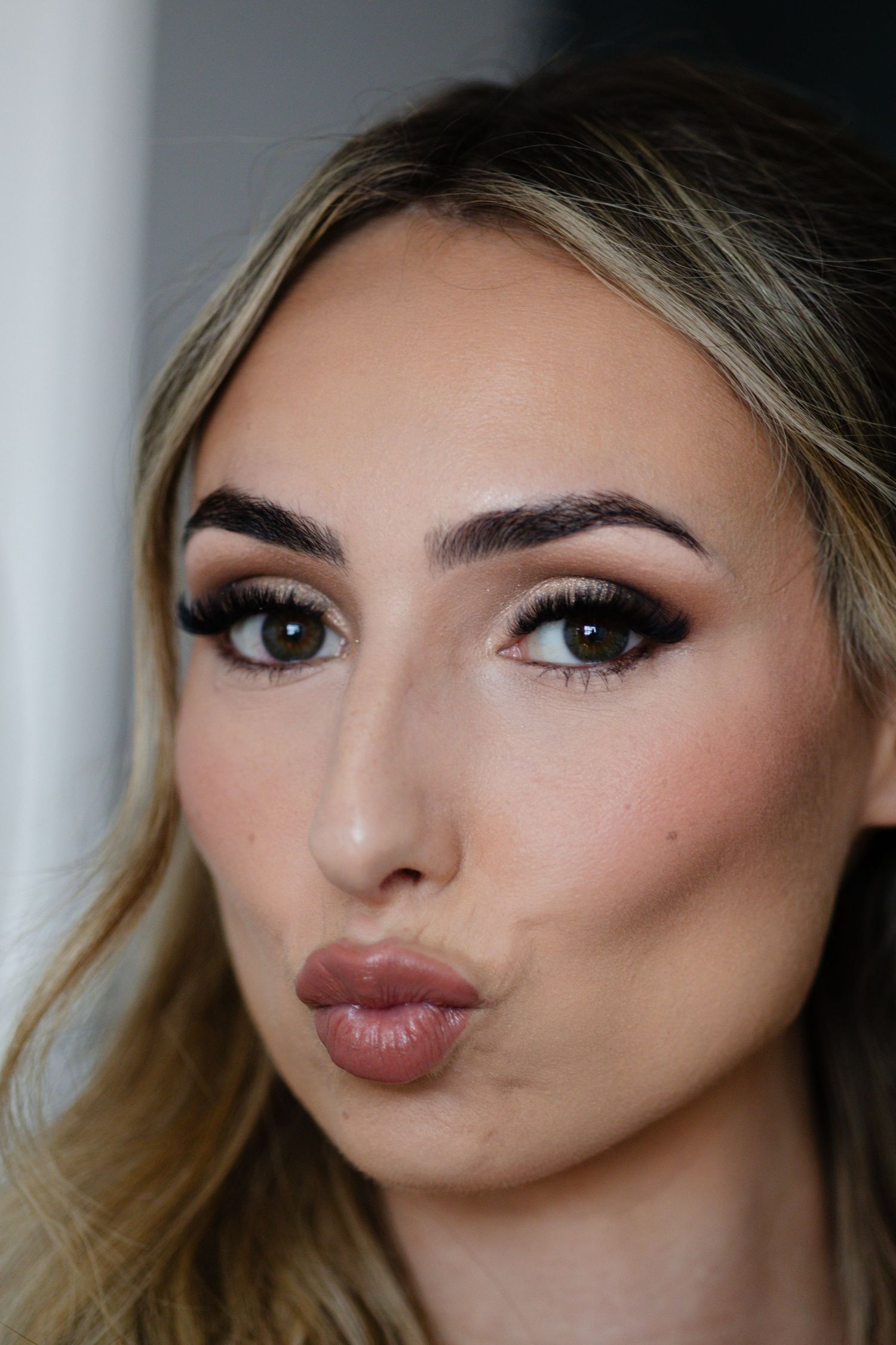 Close-up of a blonde woman with bold brows, dramatic lashes, and nude lips making a kissing face.