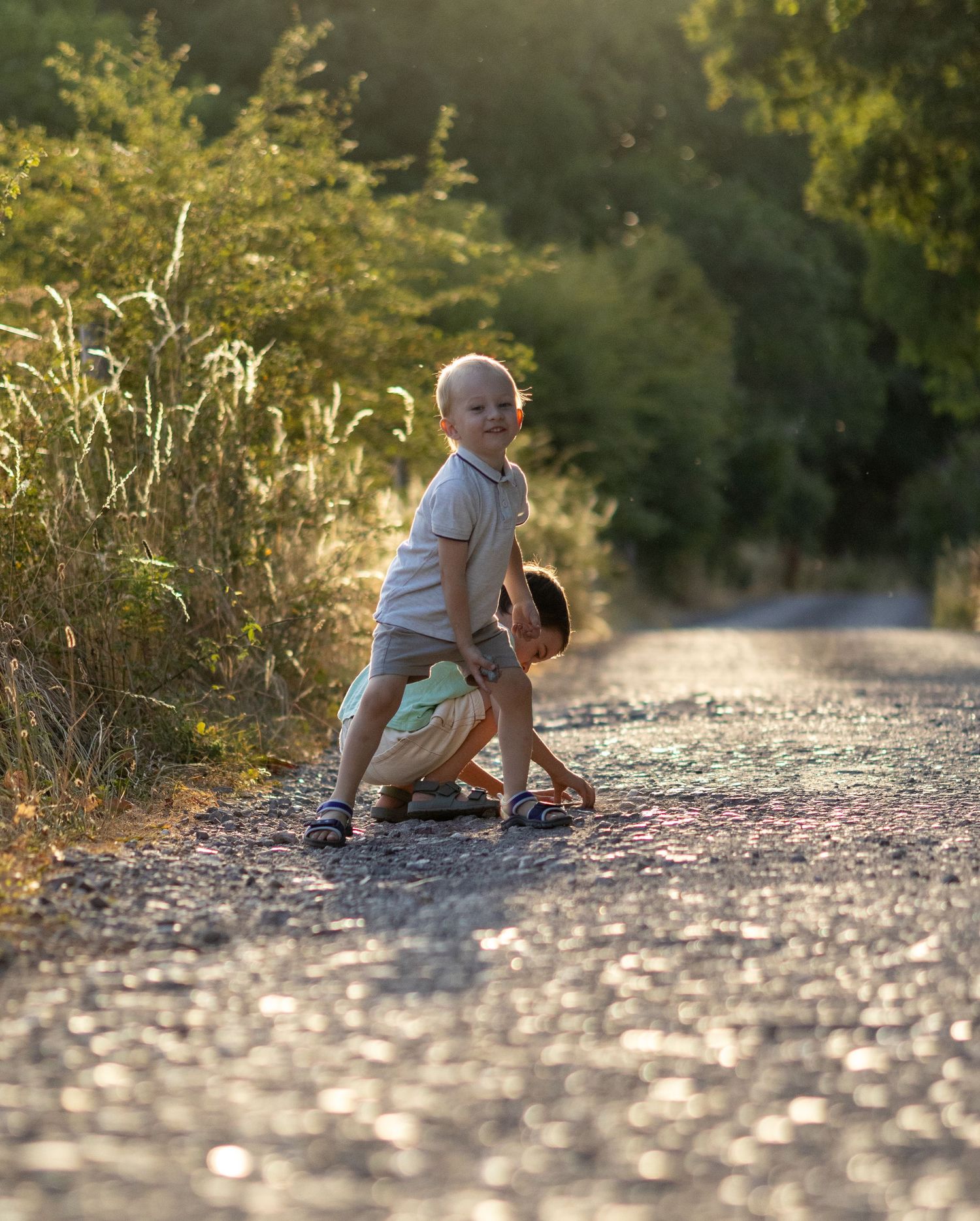 Two young boys playing on a gravel road surrounded by golden sunlight and green trees.
