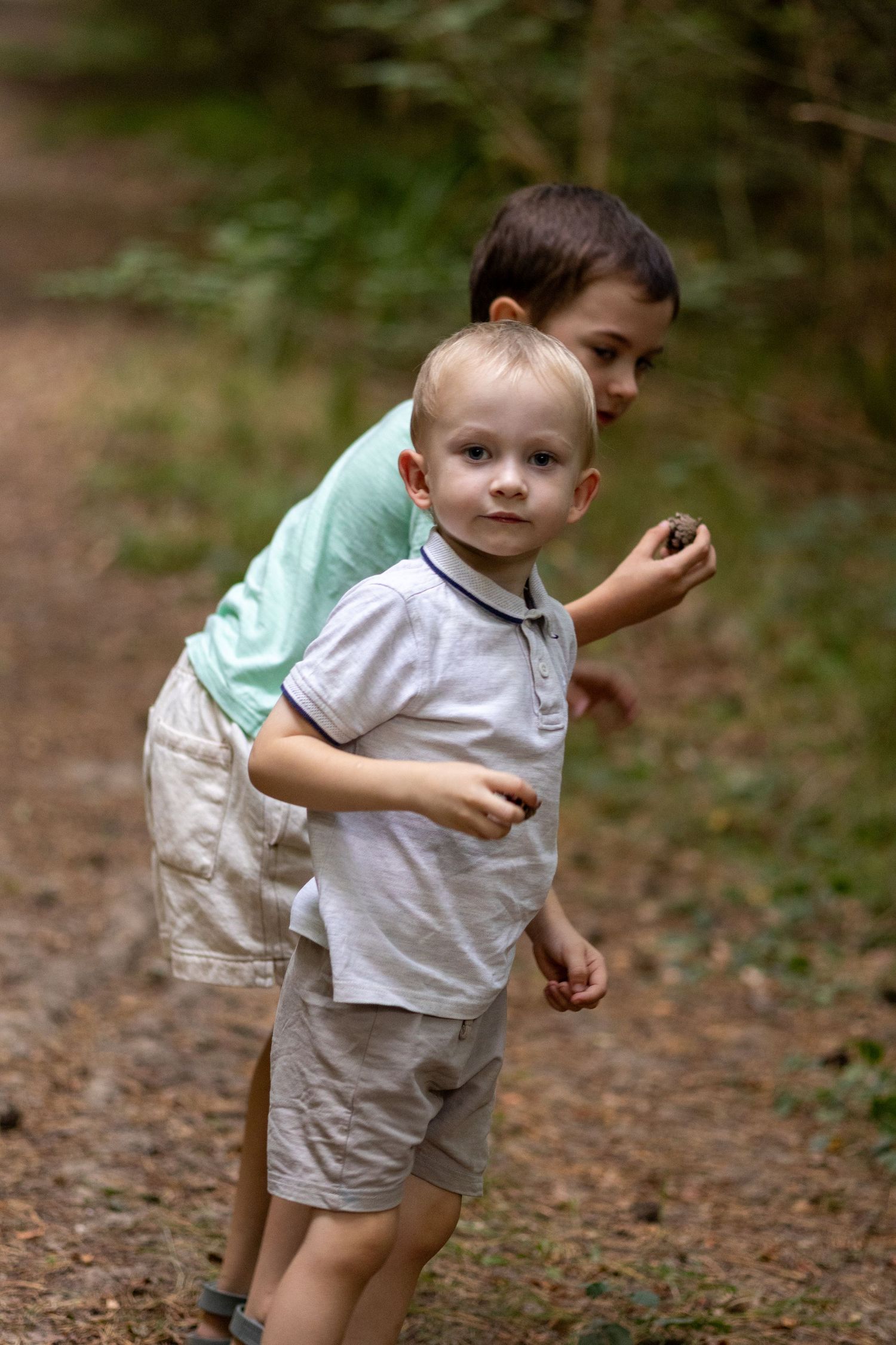 An older brother hugs his younger toddler sibling from behind on a shaded forest path.