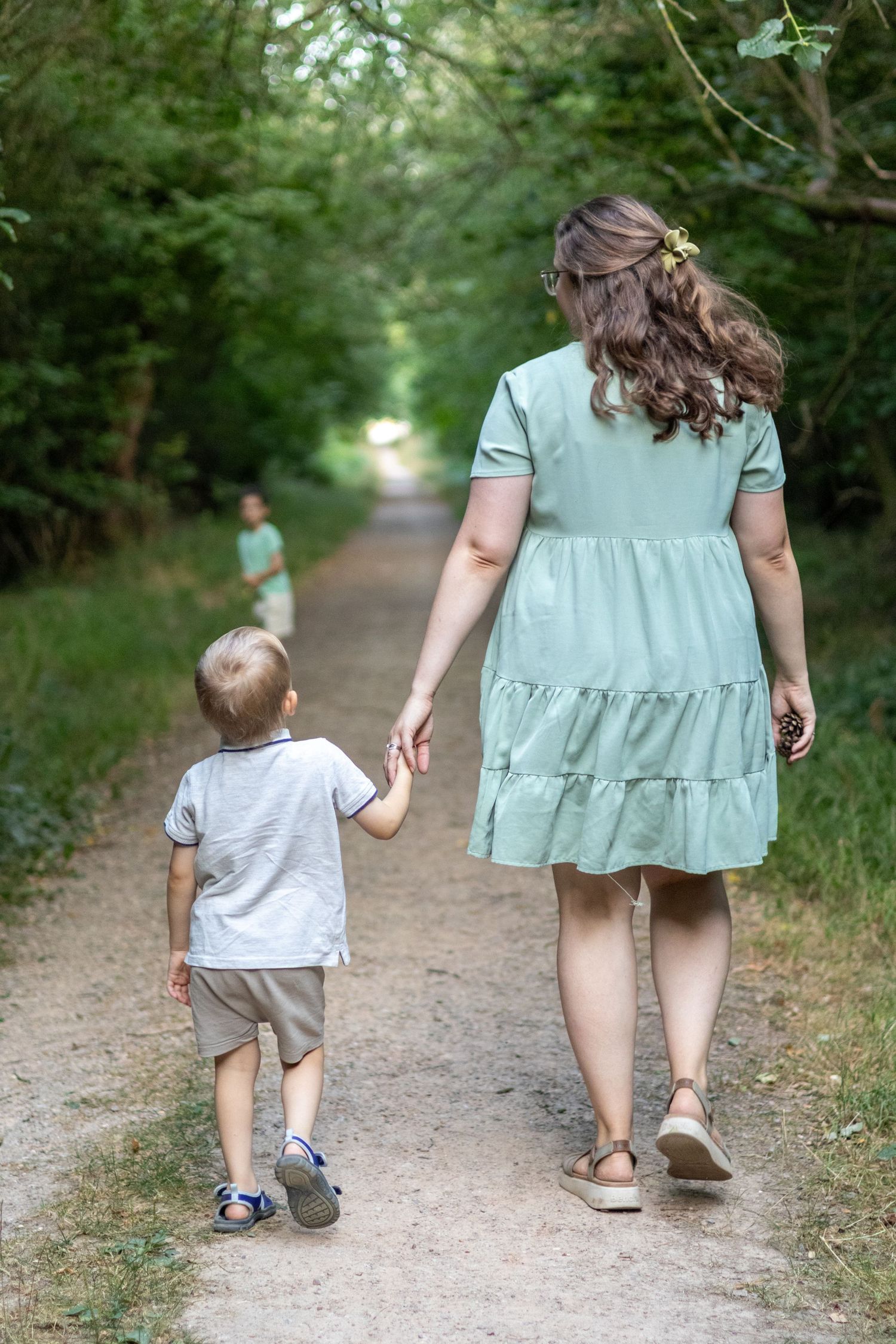 A mother in a mint dress holds her toddler son's hand while walking on a forest path.