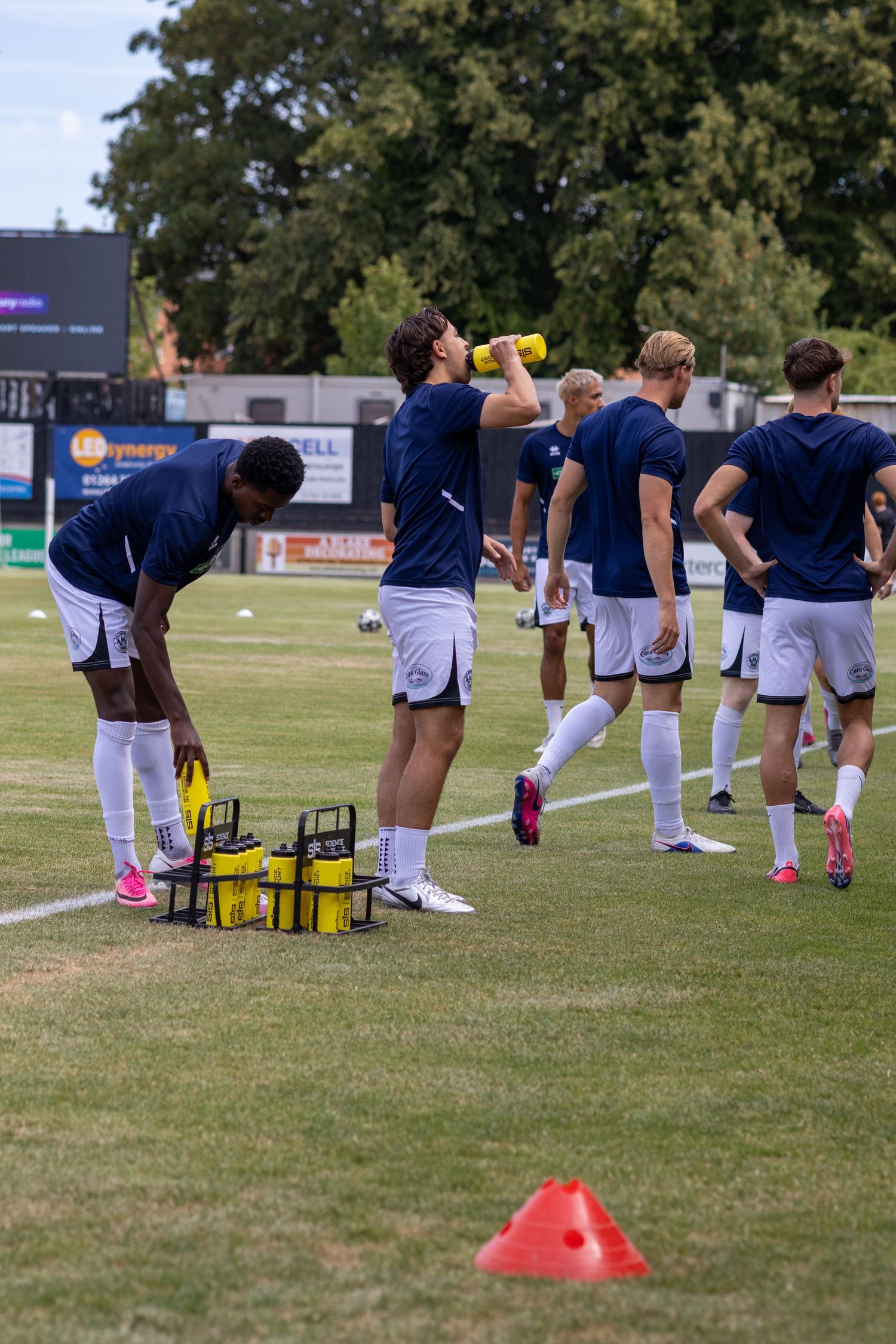 Football players in navy kits take a water break during pre-match warmup on a sunny day at a non-league ground.