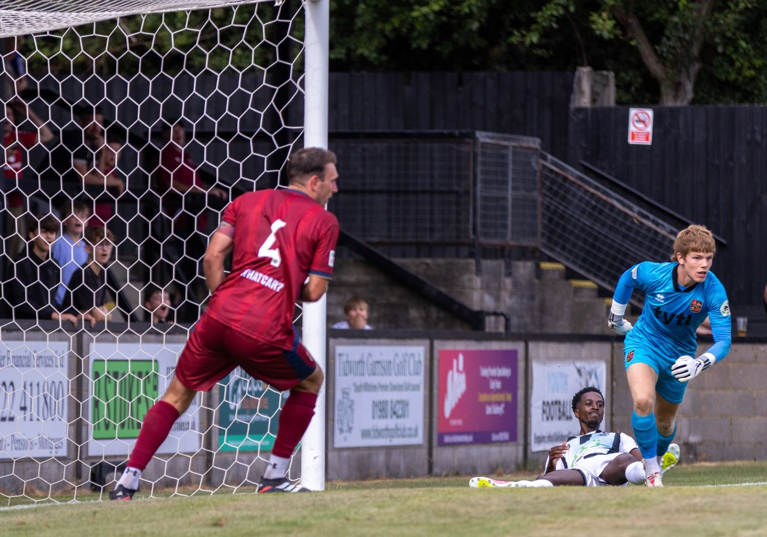 A goalkeeper reacts as a striker scores near the post during a non-league football match on a warm day.