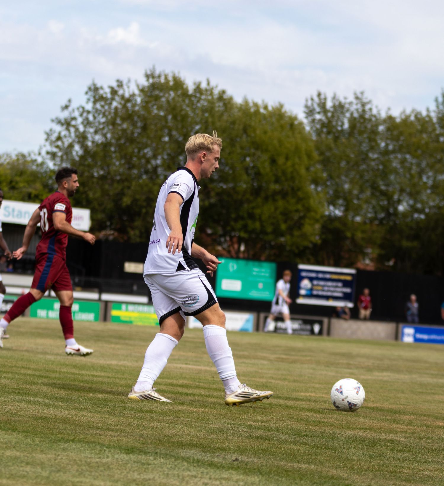 Blonde footballer in white kit controls ball during match against maroon-shirted opponents outdoors.