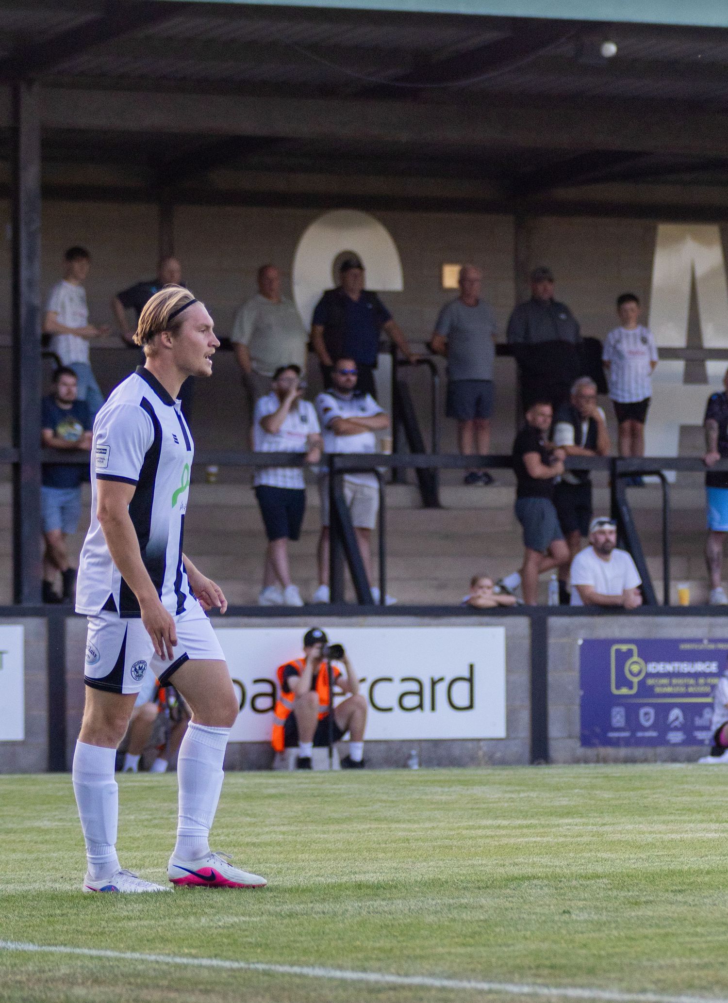 Football player in white strip stands on pitch with spectators watching from covered stand.