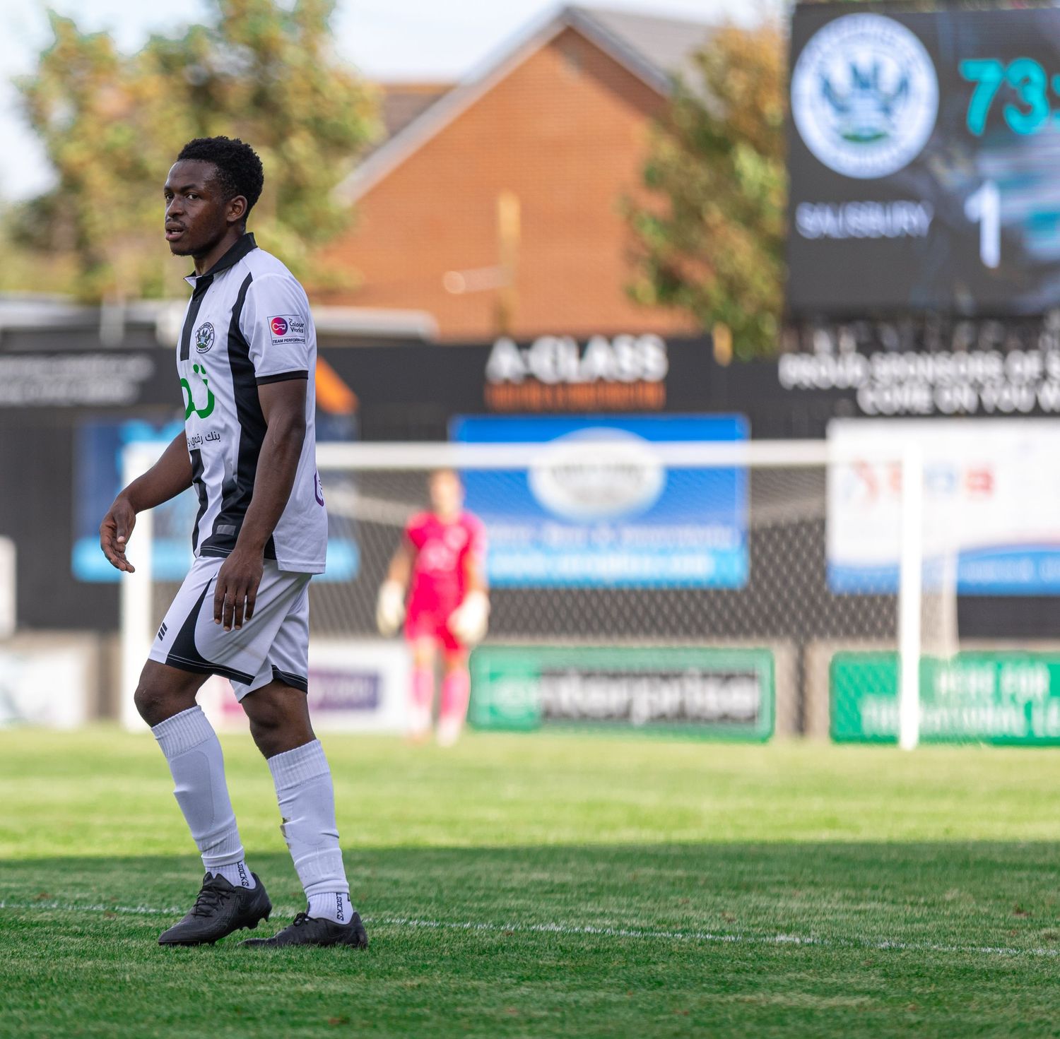 A soccer player in black and white uniform walks on the field during a match with a scoreboard showing 1-0.