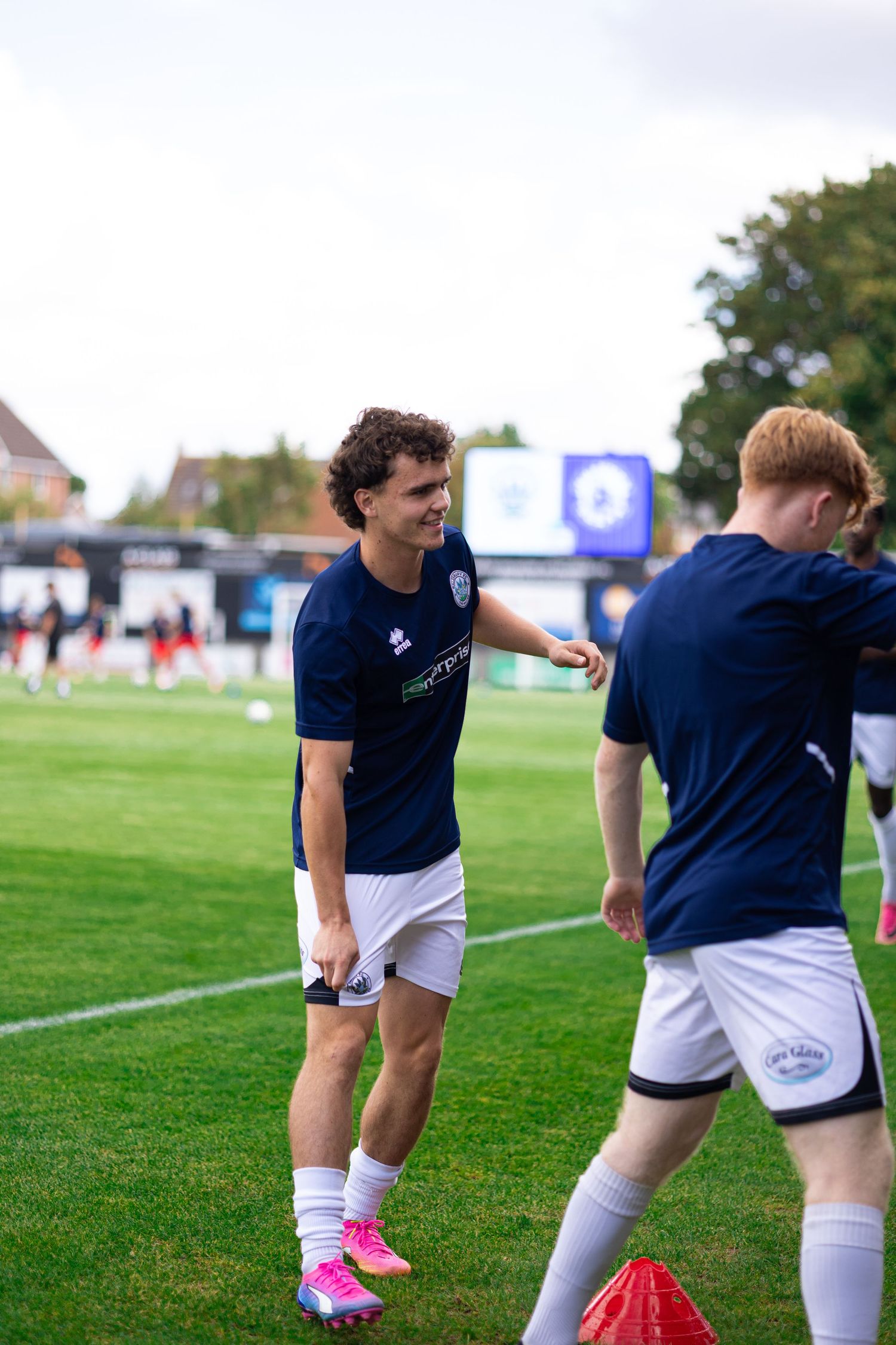 Two soccer players in navy uniforms warm up on a green field before a match, with teammates visible in the background.