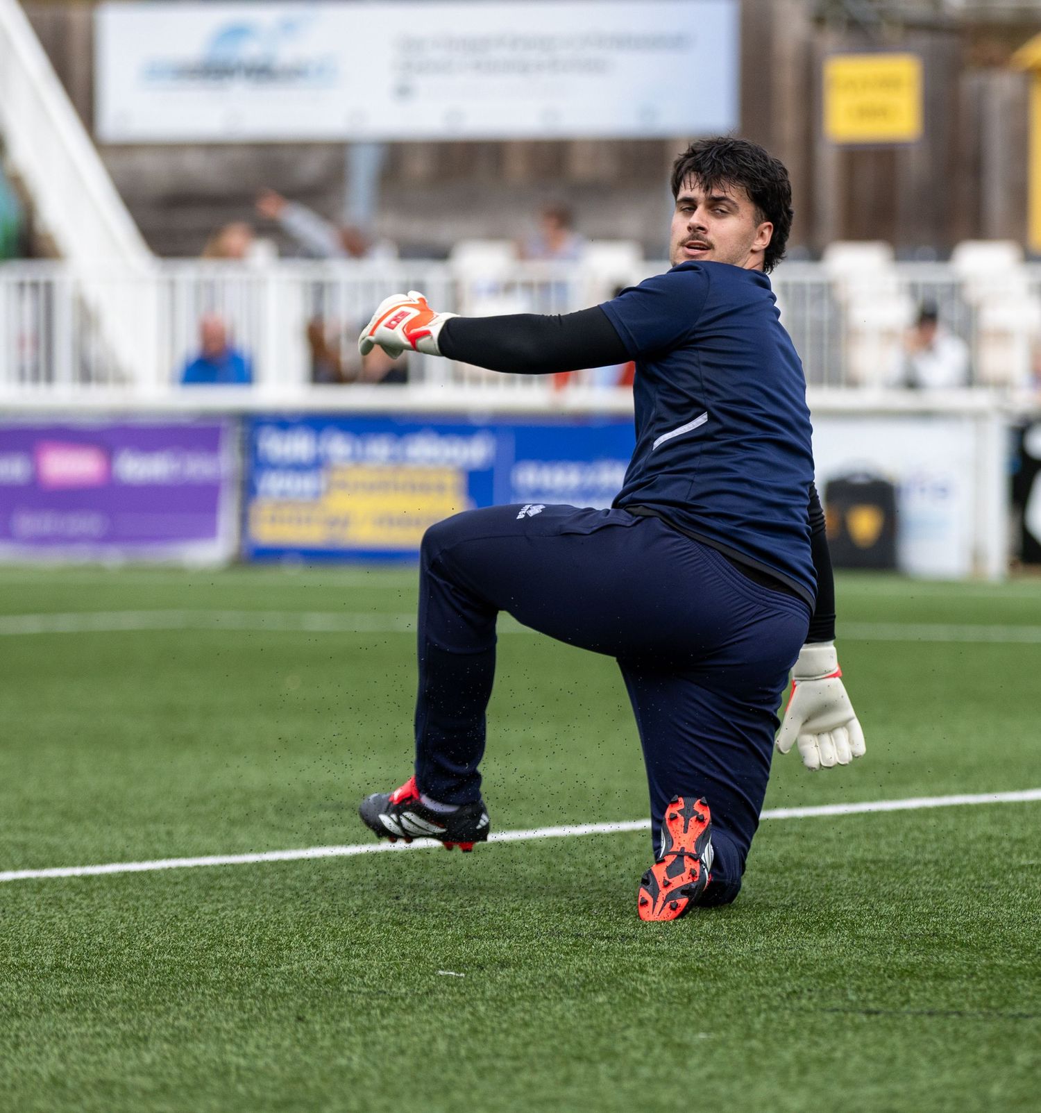 Goalkeeper in navy kit stretches to catch a ball during warm-up on an artificial turf football pitch.