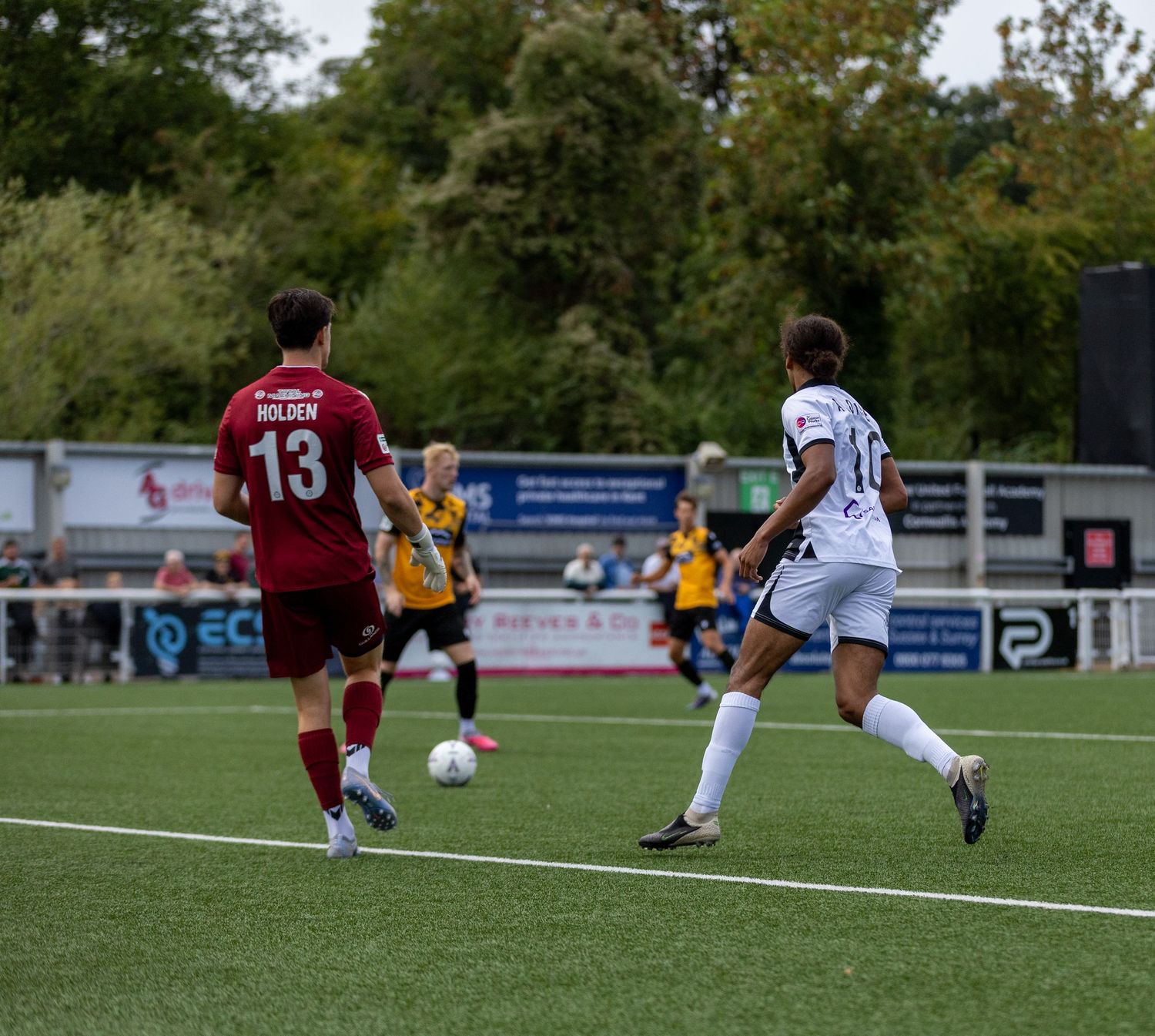 Soccer players in action during a match, player number 13 in red dribbling against opponent in white on green pitch.