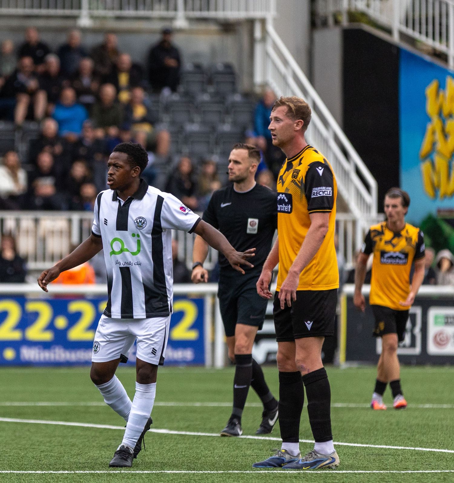 Soccer players and referee on field during a competitive match, with stadium seating visible in the background.