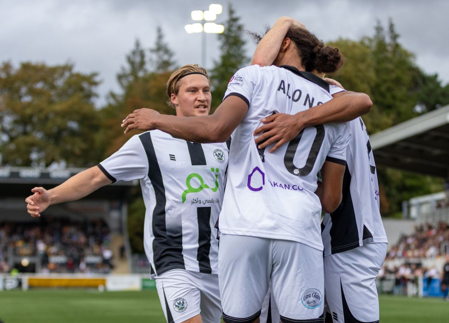 Football players in black and white jerseys celebrate a goal together on a sunny match day.