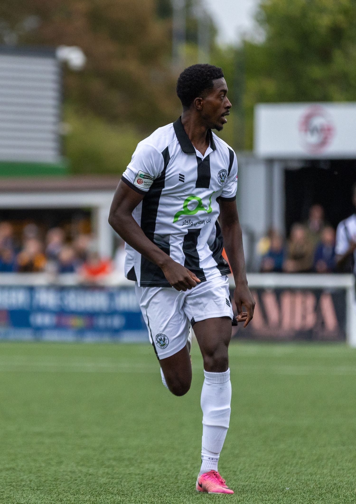 A football player in a black and white kit with pink boots runs during a match at a non-league ground.