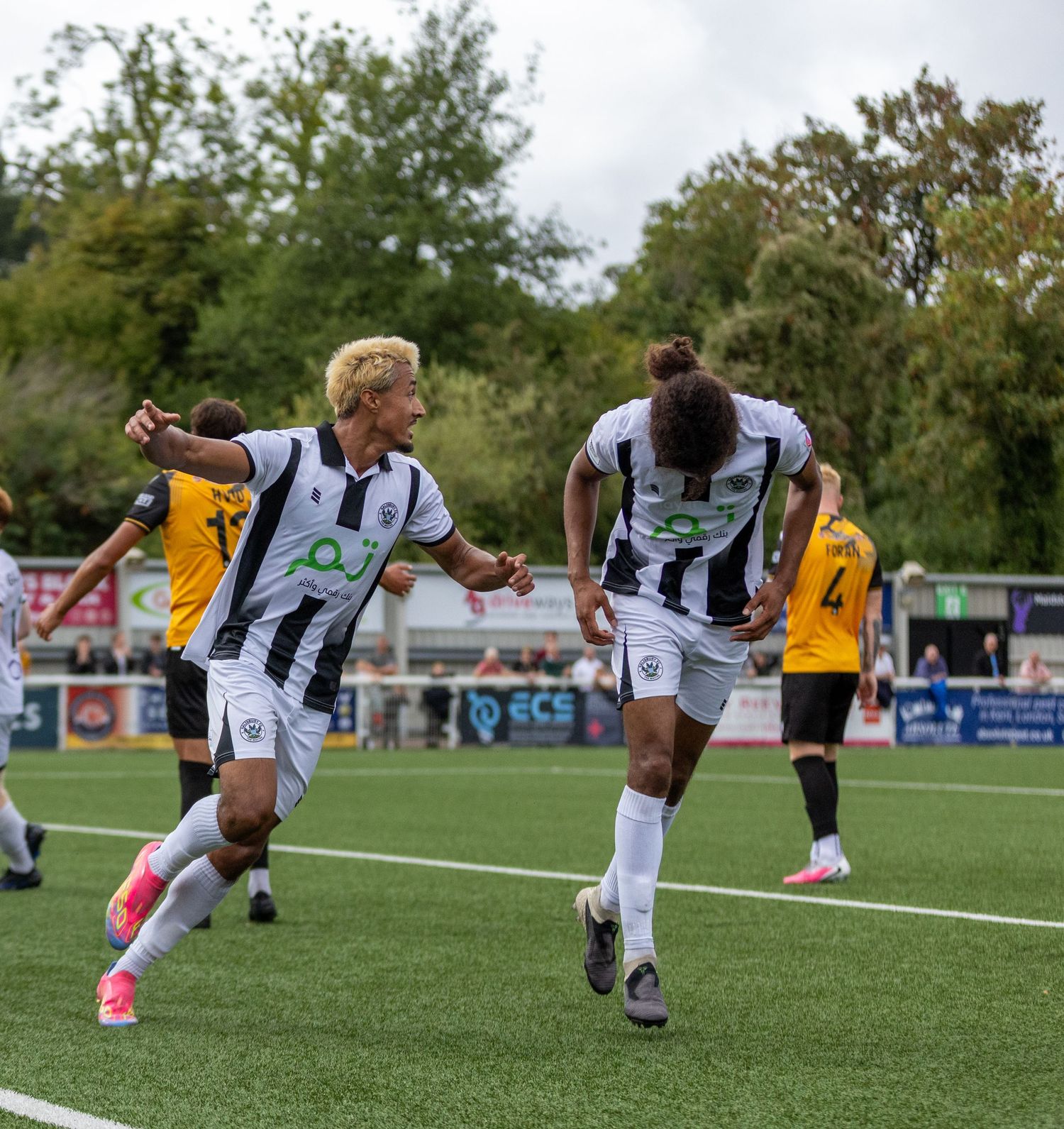Two soccer players in black and white uniforms celebrate a goal on a green pitch during a sunny outdoor match.