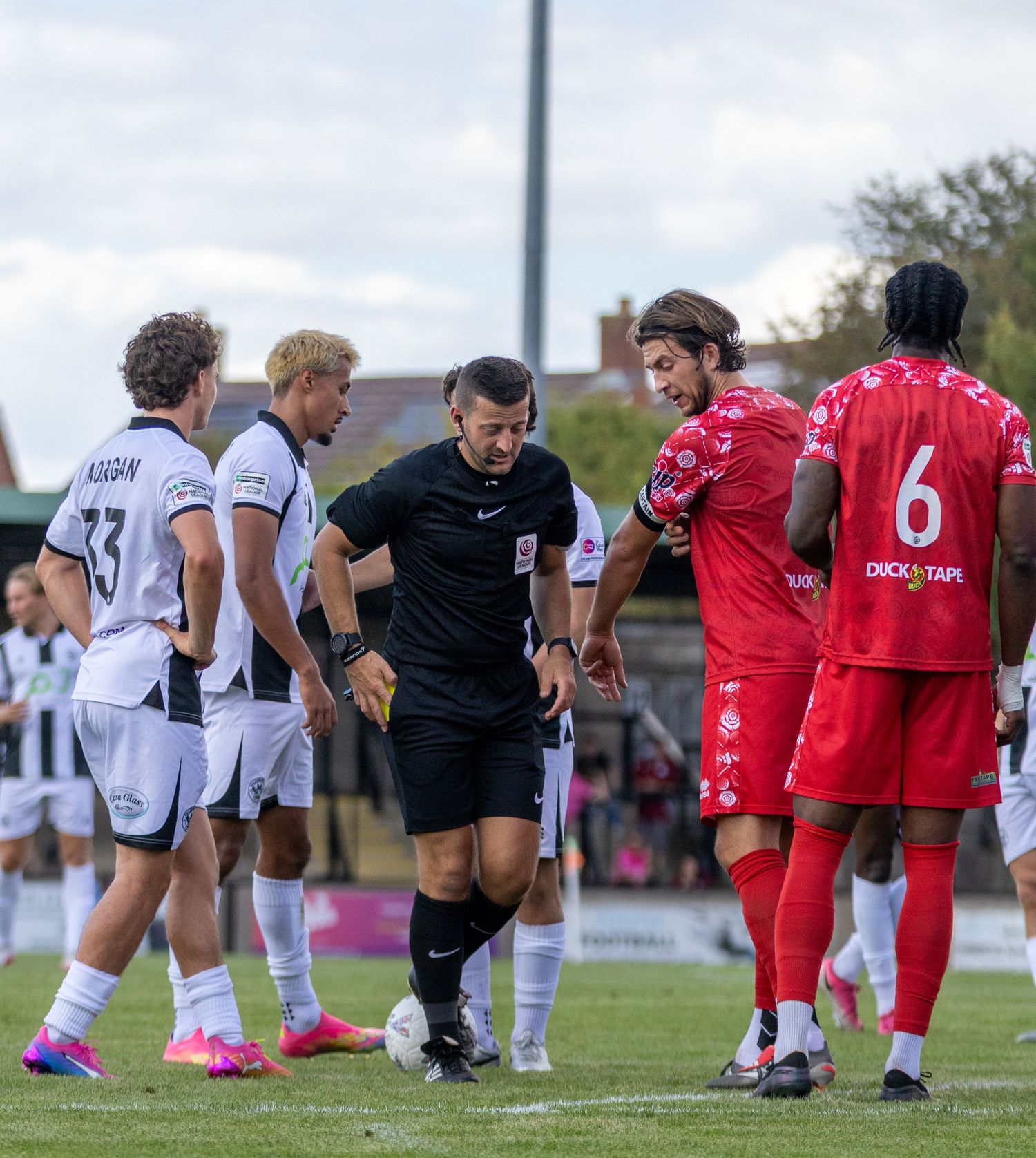 Referee and players from both teams interact during a break in play at a non-league football match.