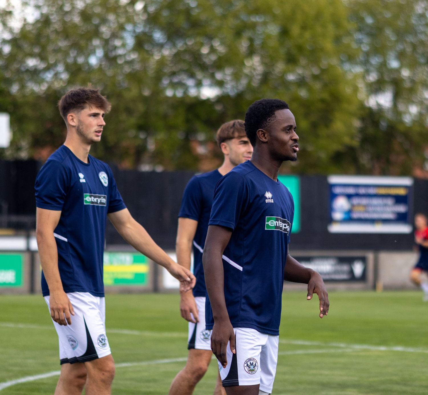 Three football players in navy blue Errea kits walk across a grass pitch during pre-match warmup.