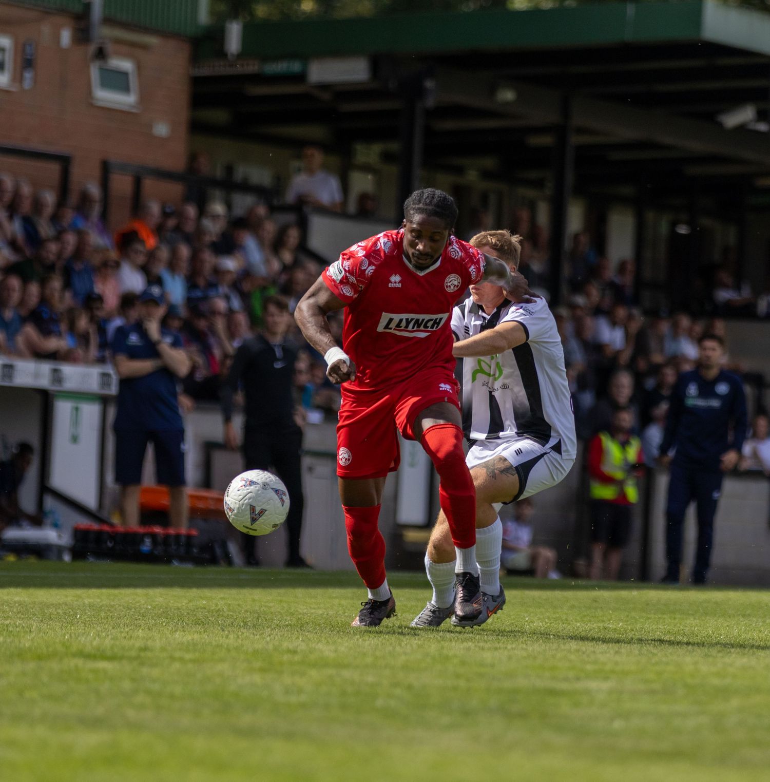 Two football players compete for possession with red attacker shielding ball from white-shirted defender.