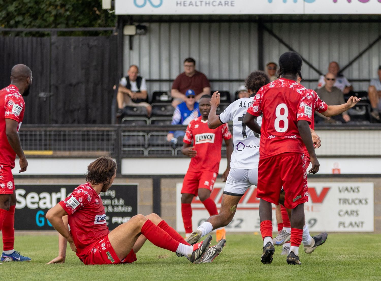 Football players in red and white kits compete during an outdoor soccer match on a sunny day.