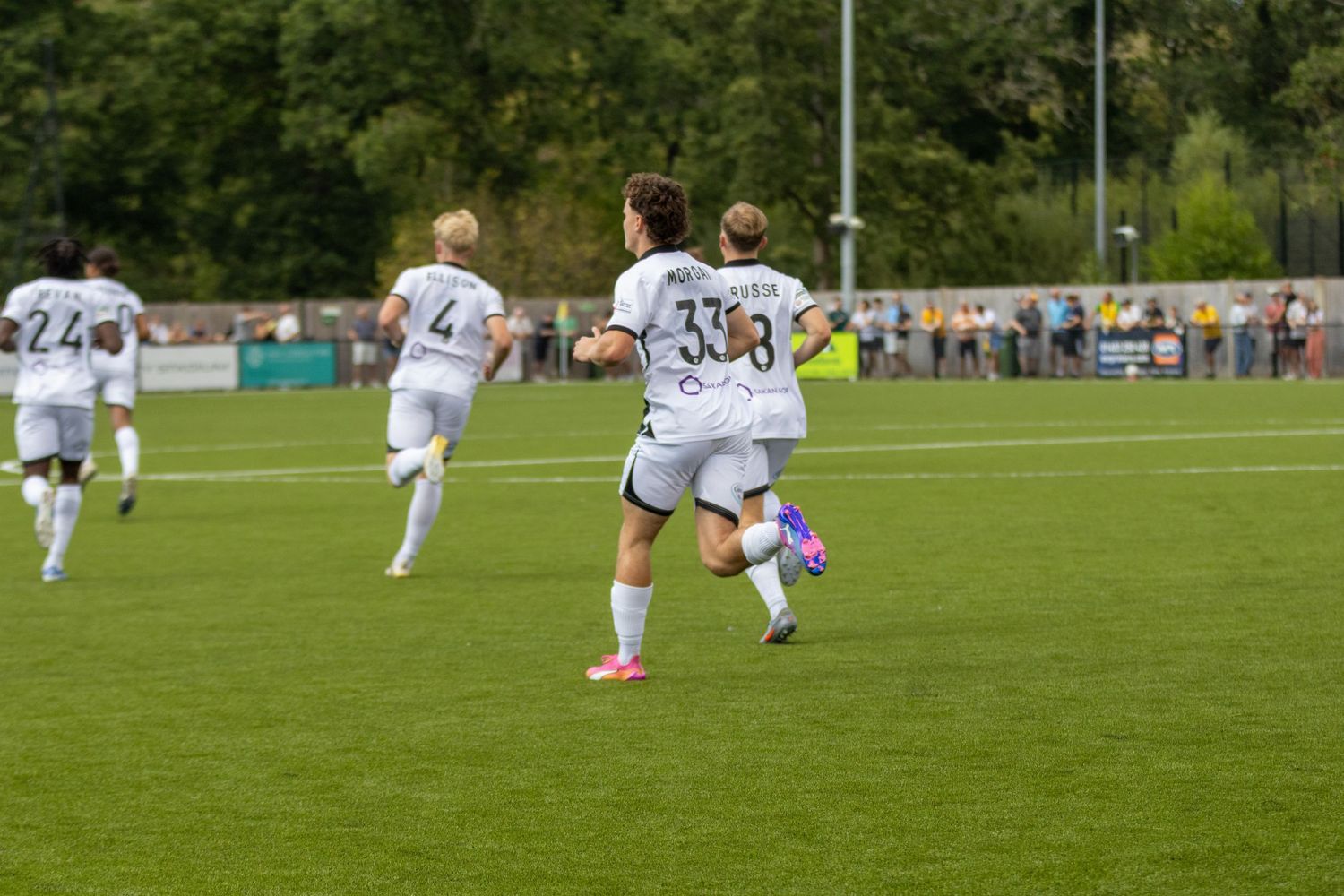 Players in white kits run across a green football pitch during a non-league soccer match on a cloudy day.