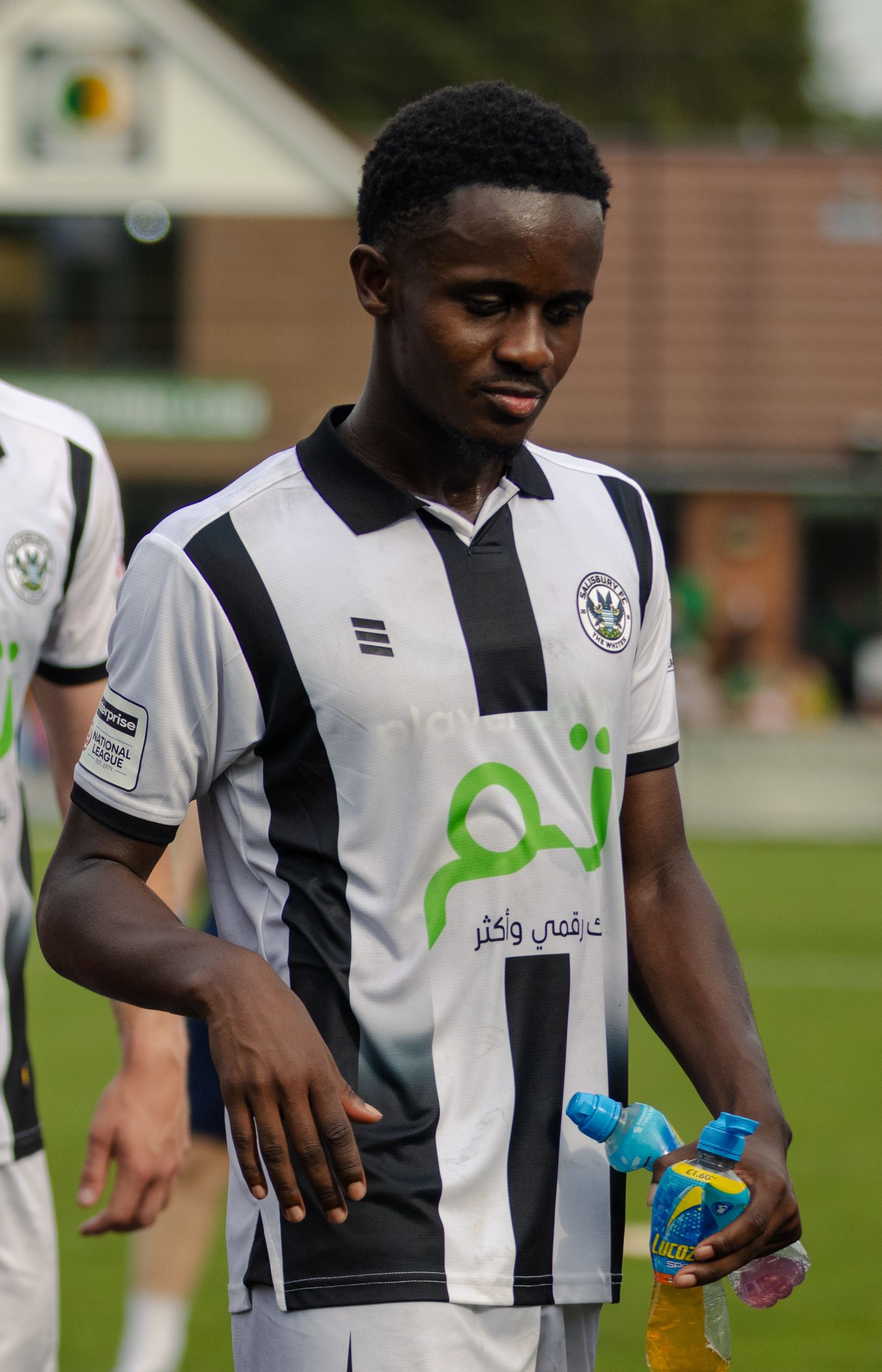 A young Black footballer in black and white striped kit holds a water bottle on a football pitch.