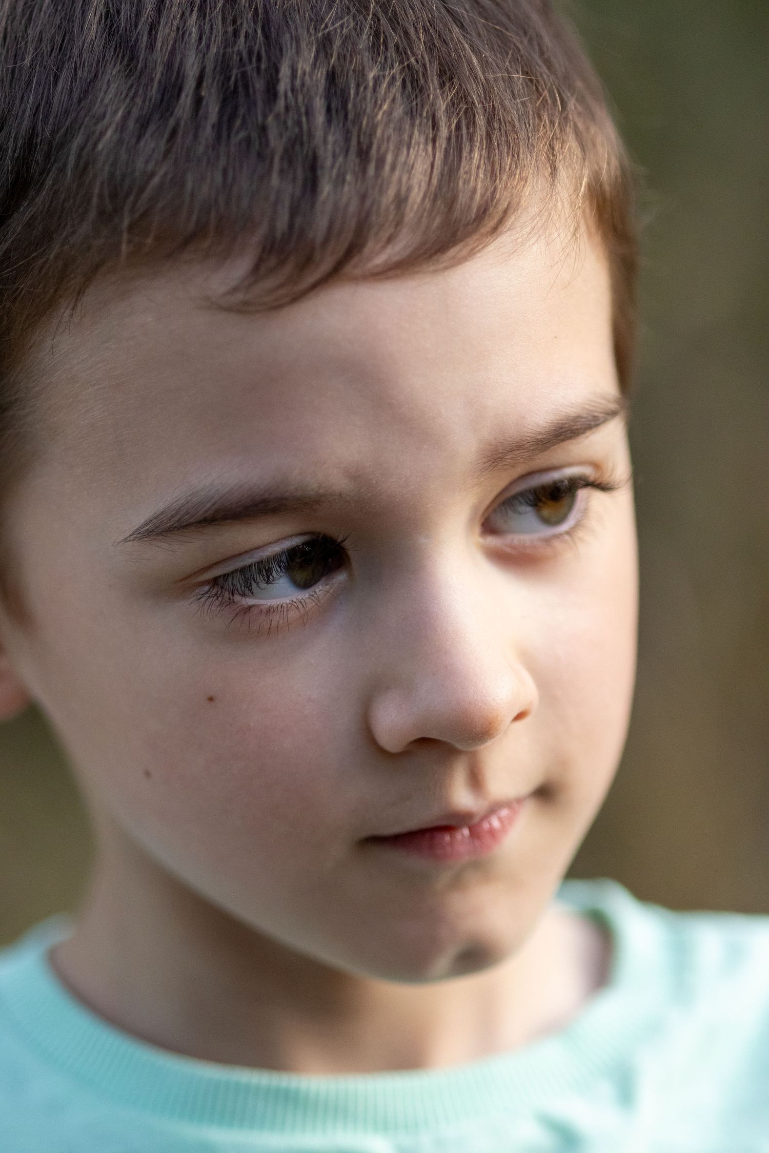 Close-up portrait of a young boy with brown eyes wearing a light blue shirt outdoors.
