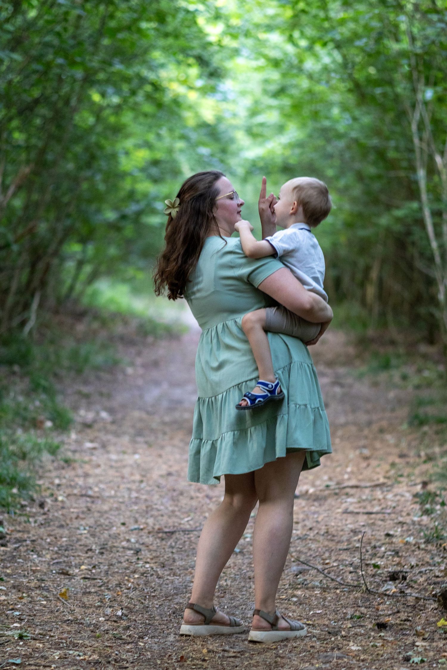 Mother holding young child on woodland path as baby points upward curiously.