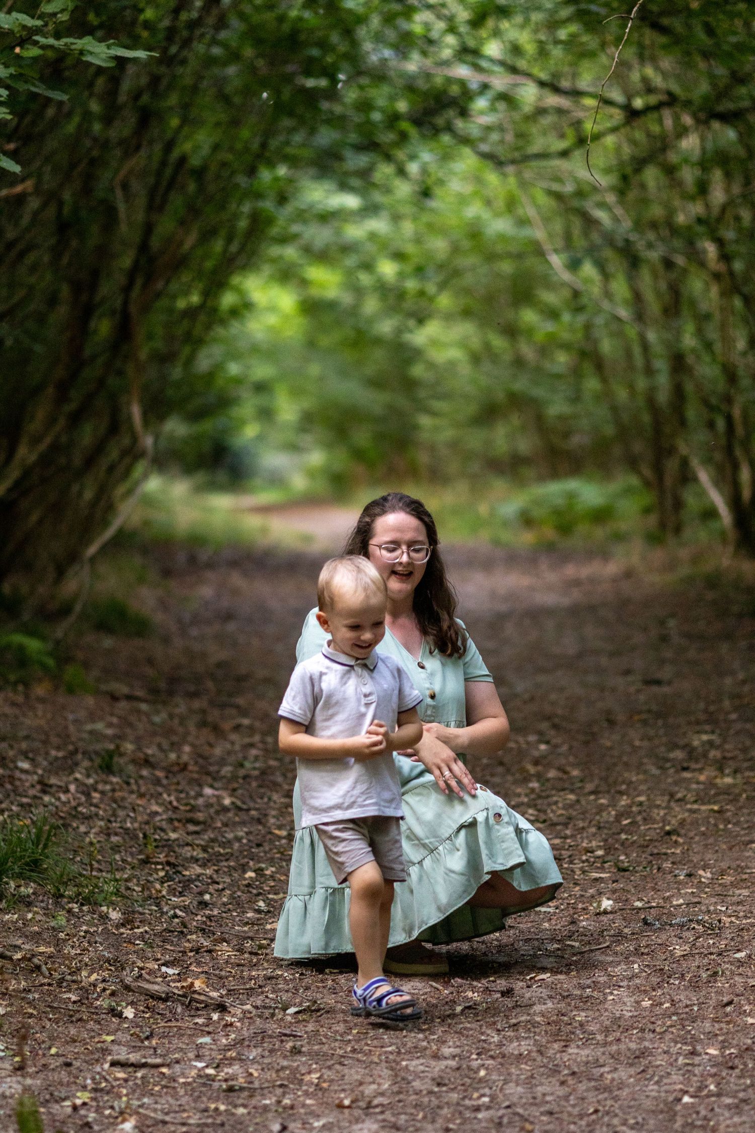 Mother crouching on forest path with toddler son in green woodland tunnel.