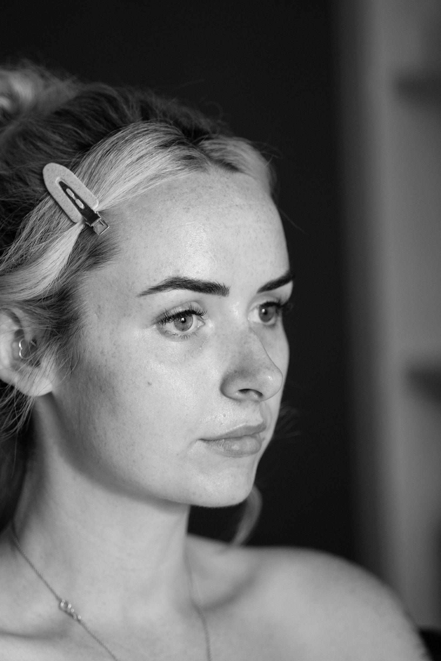 Black and white portrait of blonde woman with hair clip and delicate necklace looking away from camera.