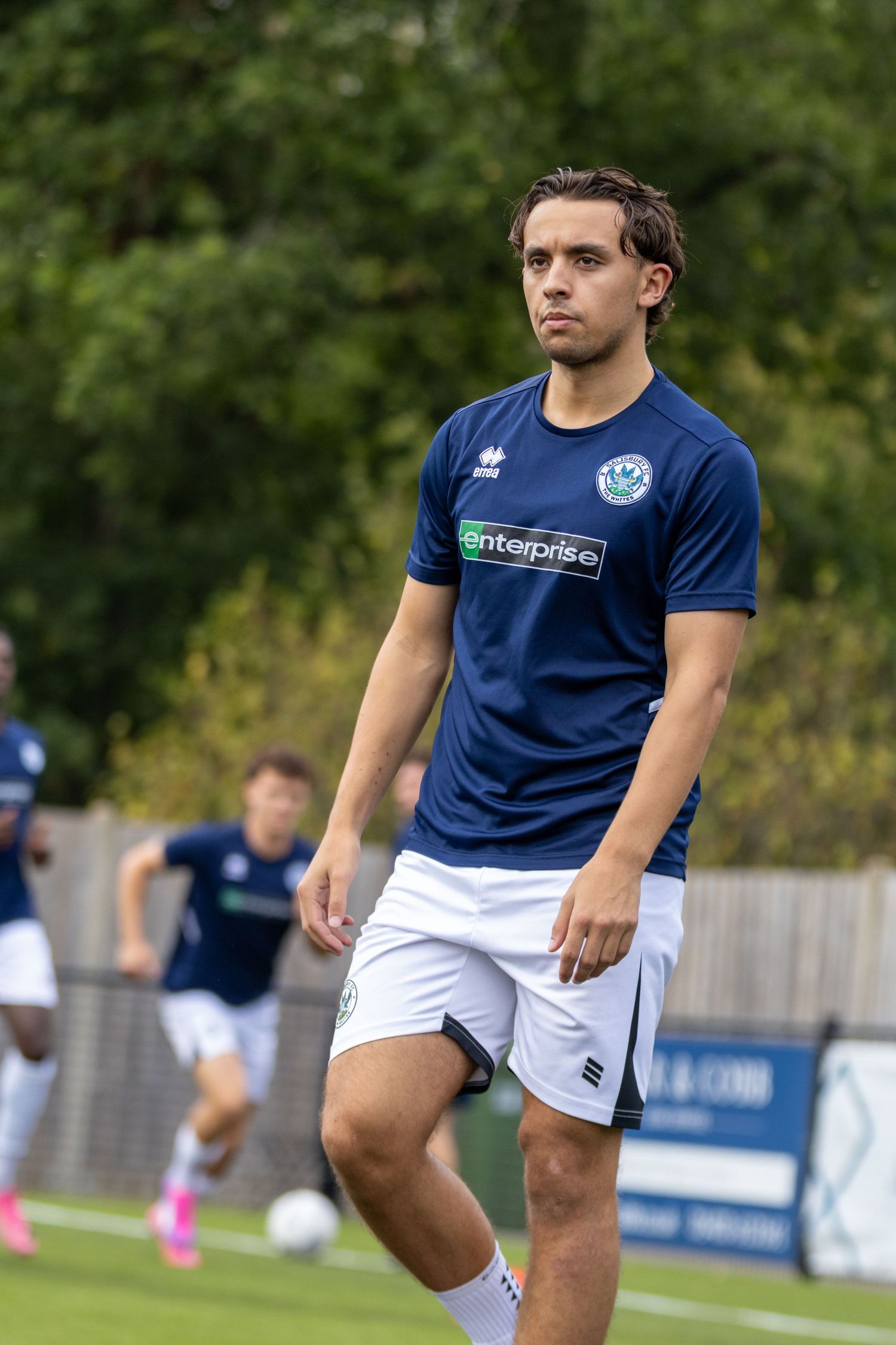 Young male footballer in navy Enterprise-sponsored kit walks across green pitch during pre-match warmup.