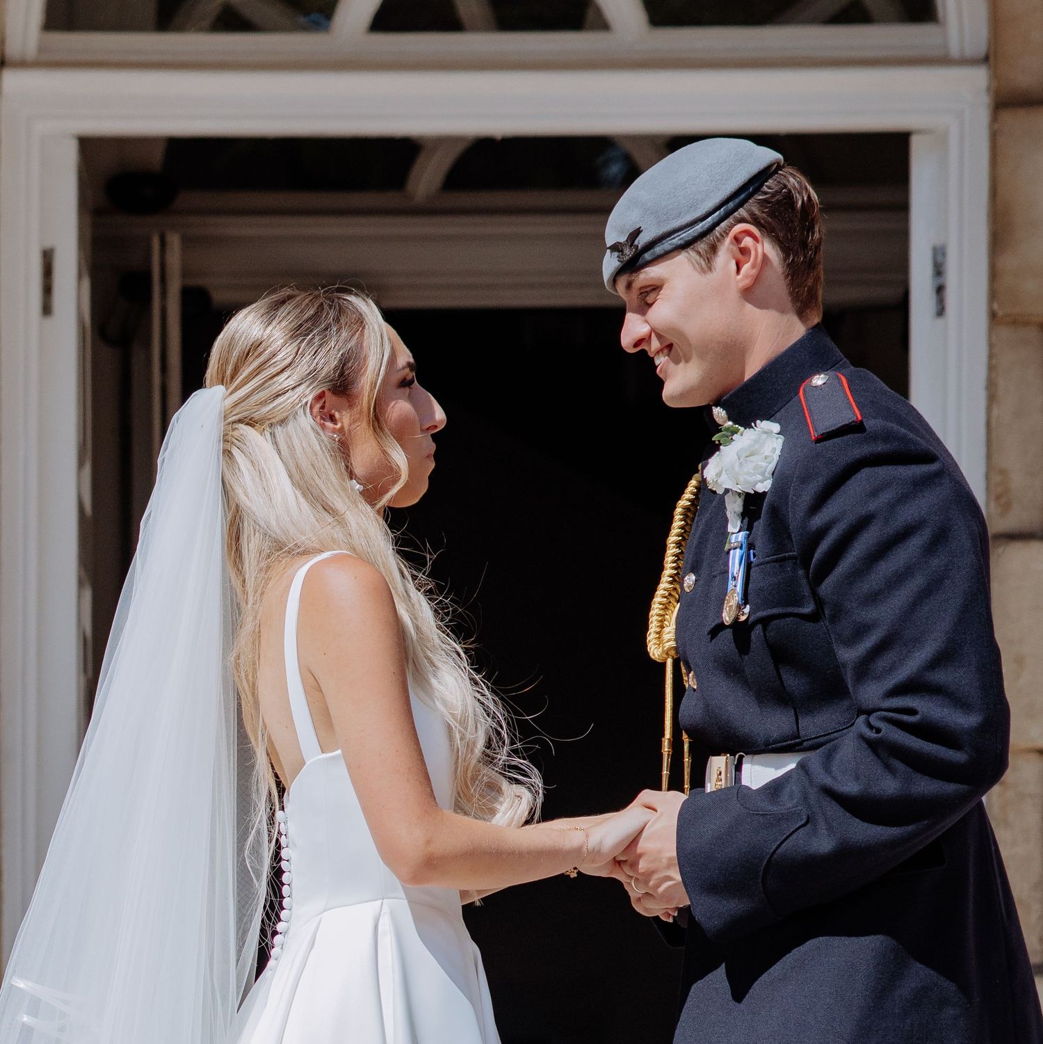 Smiling bride in white gown holds hands with groom in military dress uniform outside stone building wedding venue.