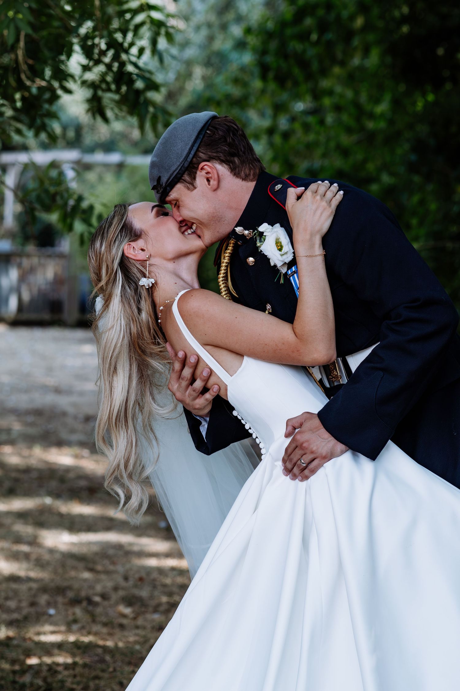 Bride in white gown and groom in military uniform sharing a romantic dip kiss at outdoor wedding.