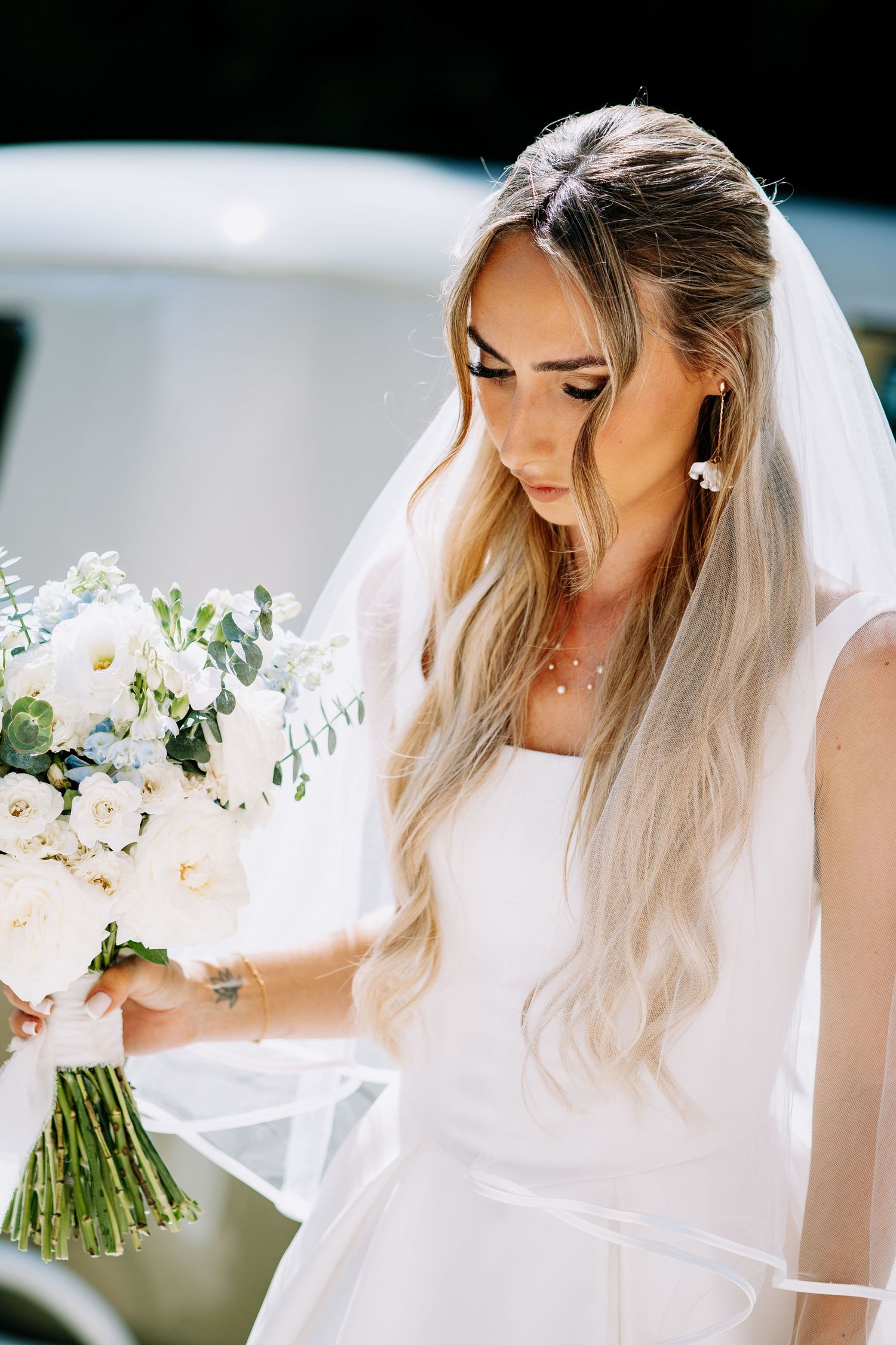Bride in sleeveless white wedding dress and veil gazing at white floral bouquet outdoors.