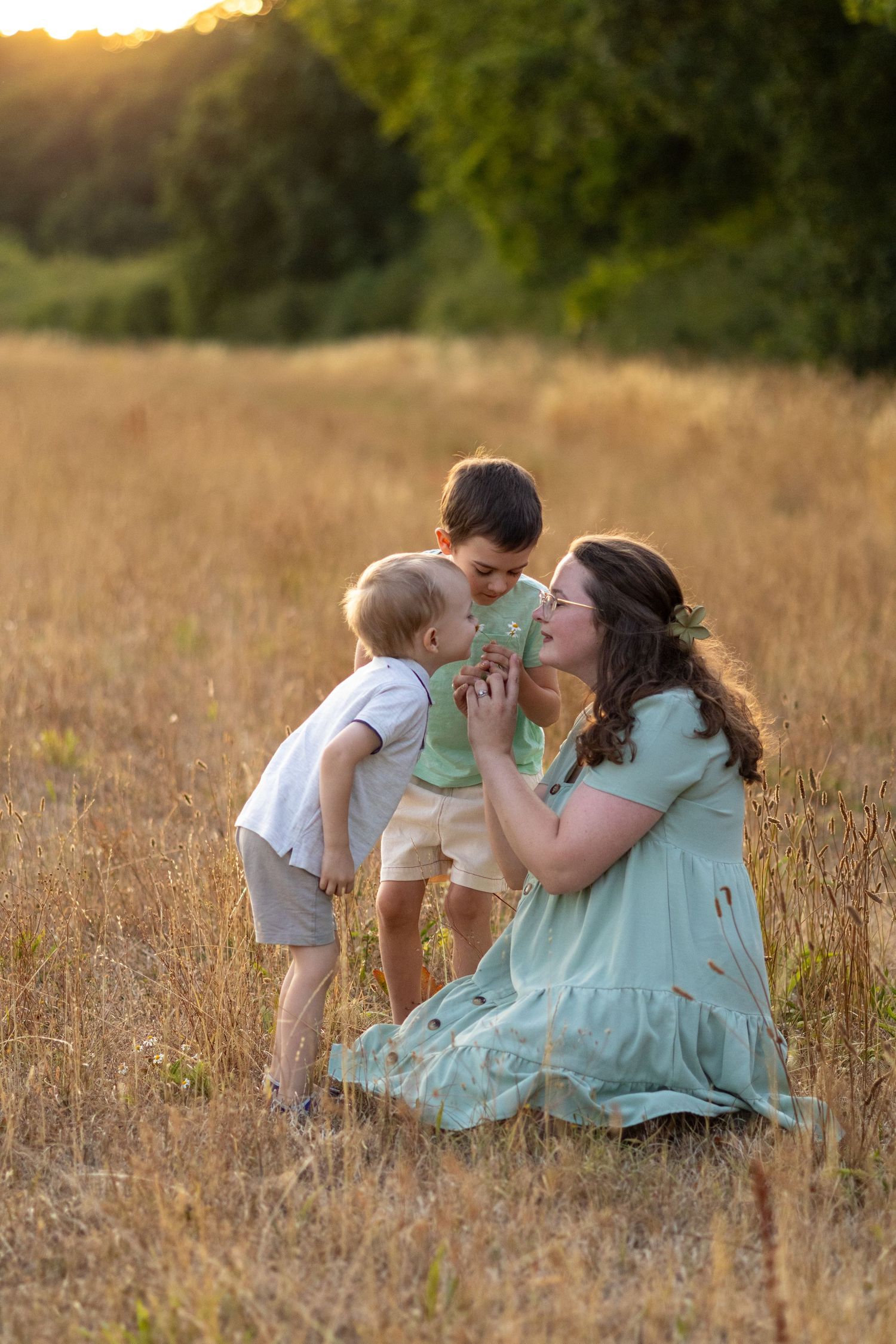 A pregnant mother in a mint dress kneels in a sunlit field while two young sons share a tender moment with her.