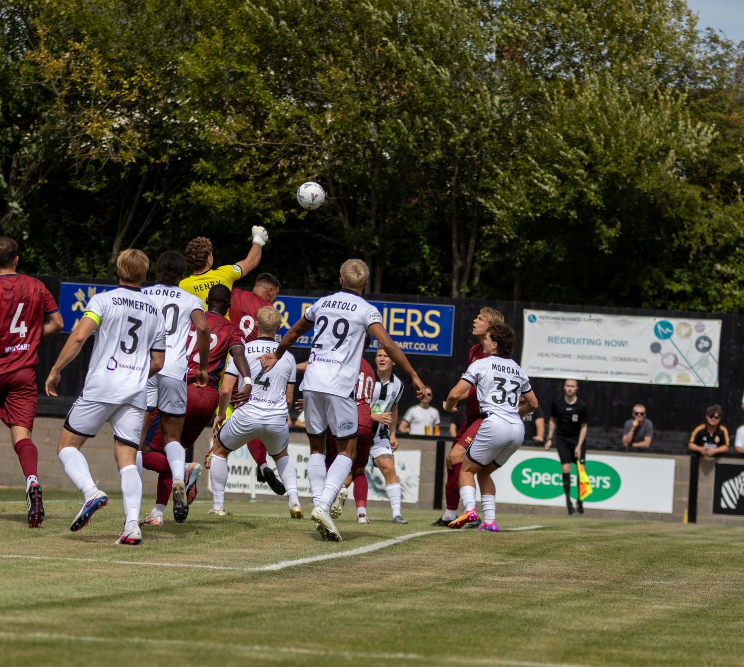 Goalkeeper punches ball clear during crowded penalty box aerial challenge in outdoor football match.