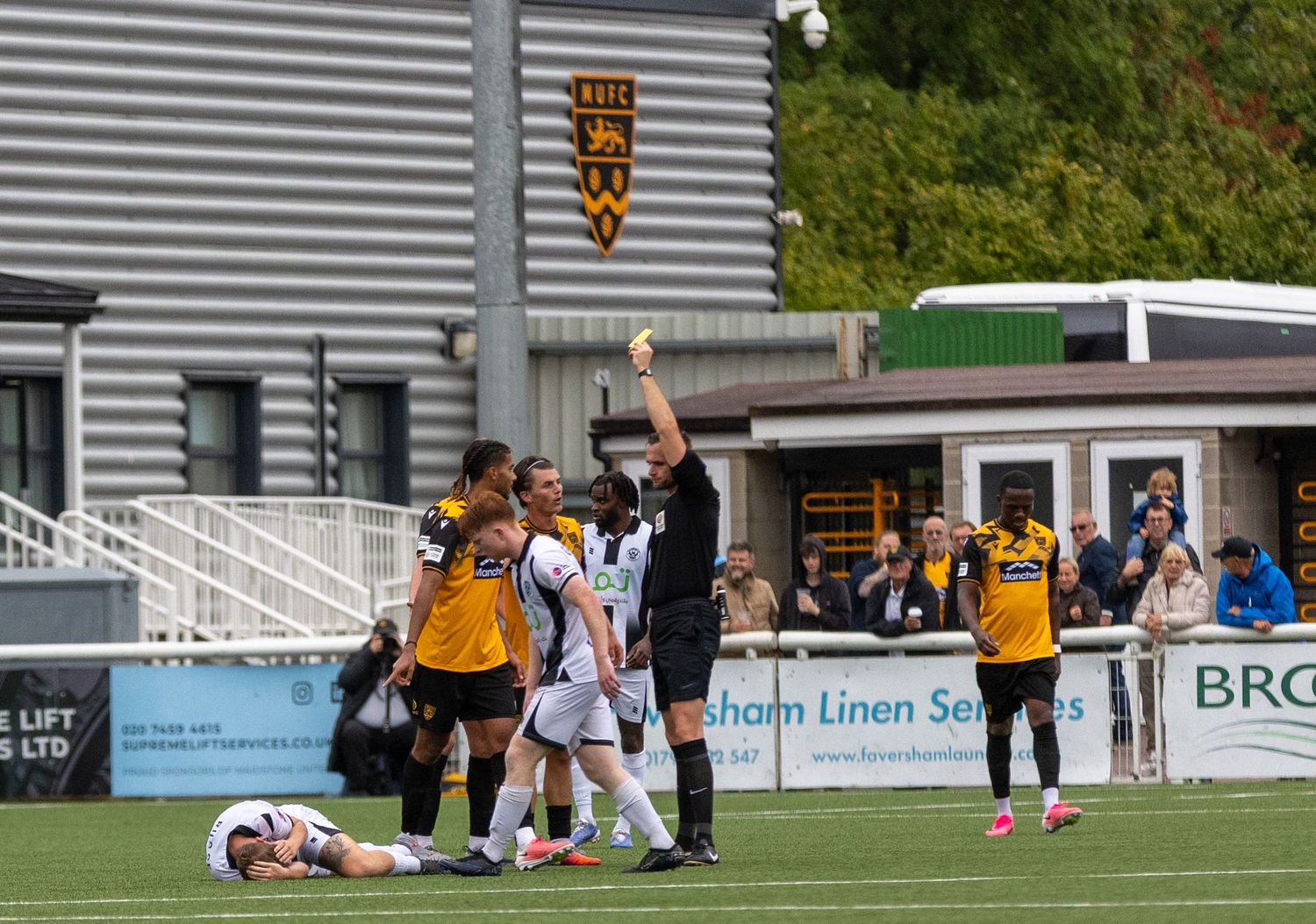 Referee shows yellow card during a soccer match, with players gathered around and one player lying on the ground.