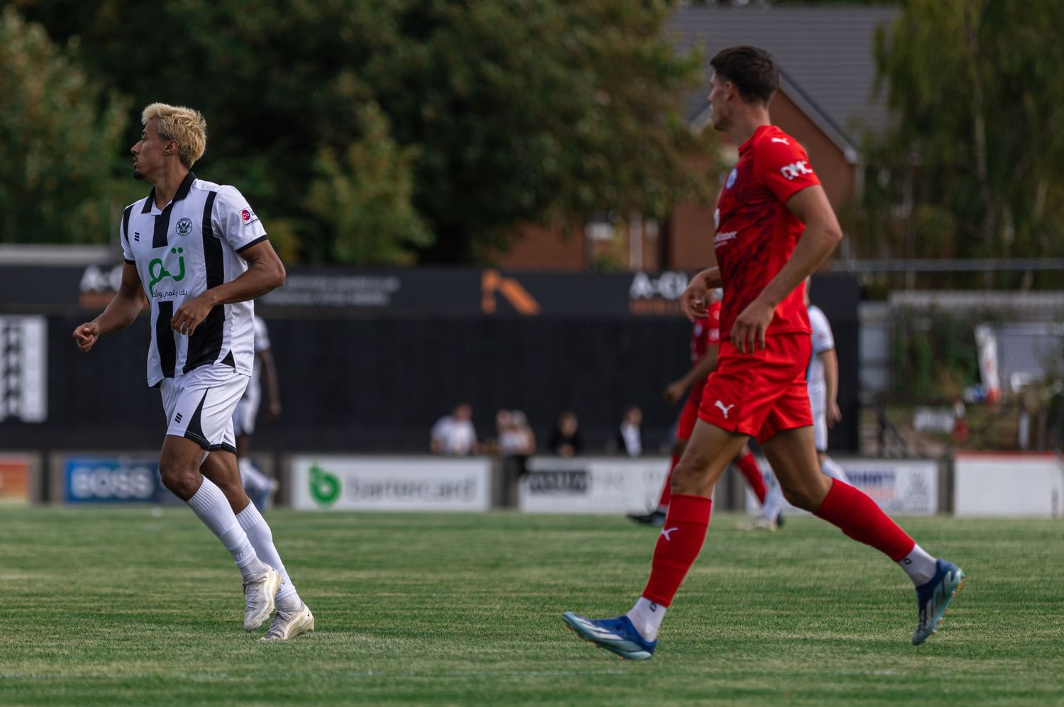 Two soccer players, one in black and white and one in red, compete on a green field during a sunny outdoor match.