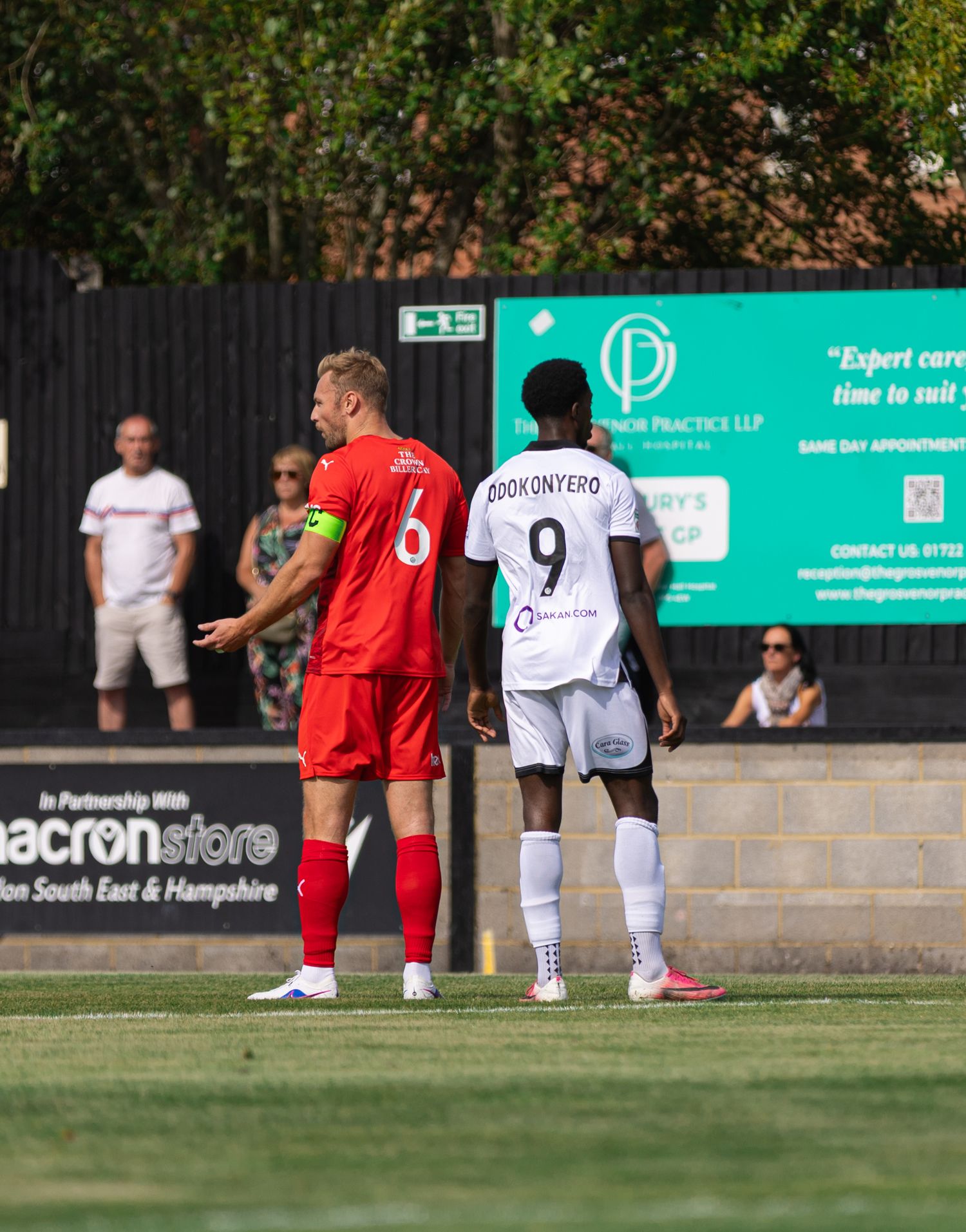 Two football players, wearing red number 6 and white number 9 jerseys, stand on a sunny grass pitch during a match.