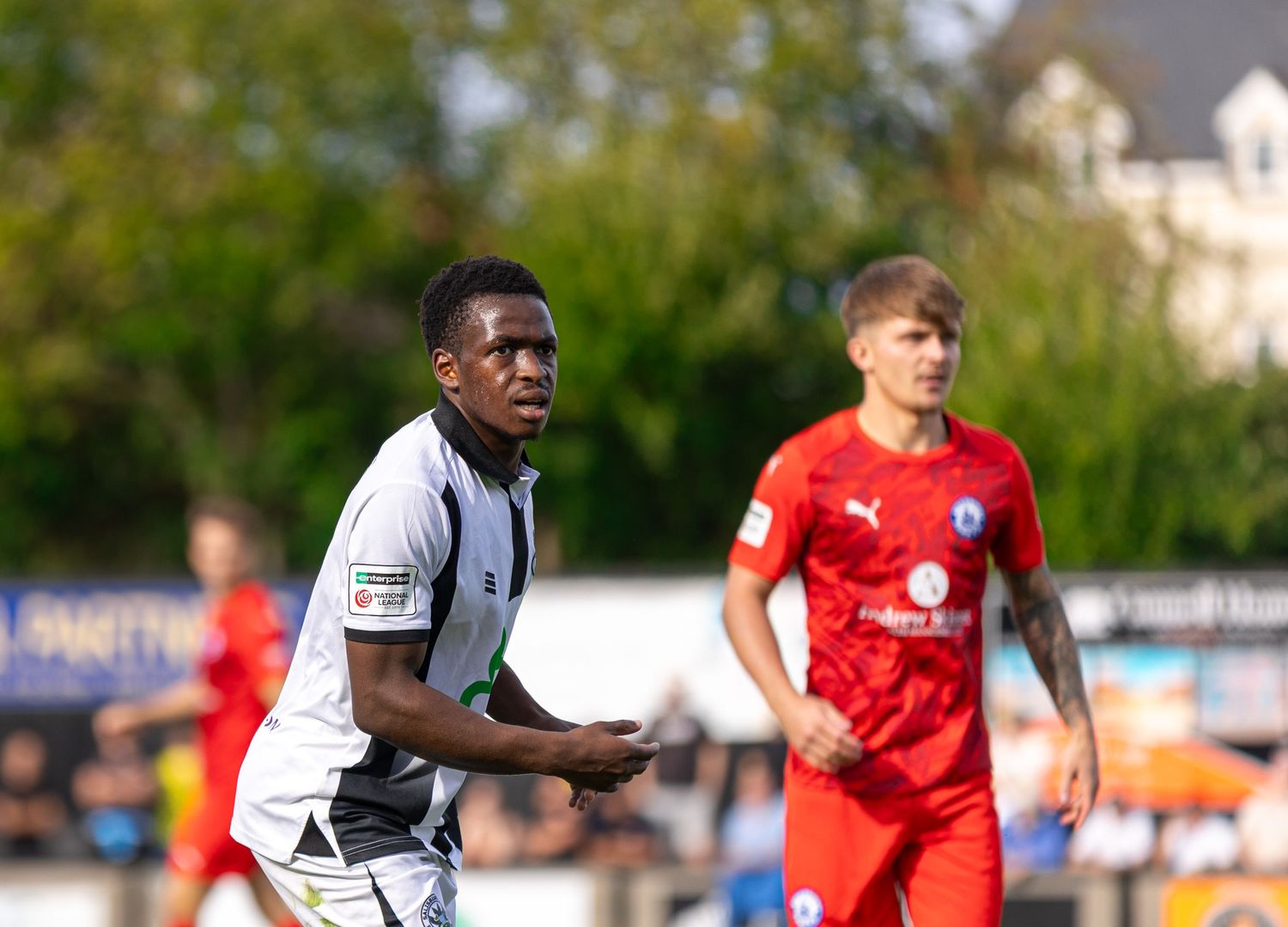Two soccer players on a sunny field, one in black kit and one in red, during a competitive match.
