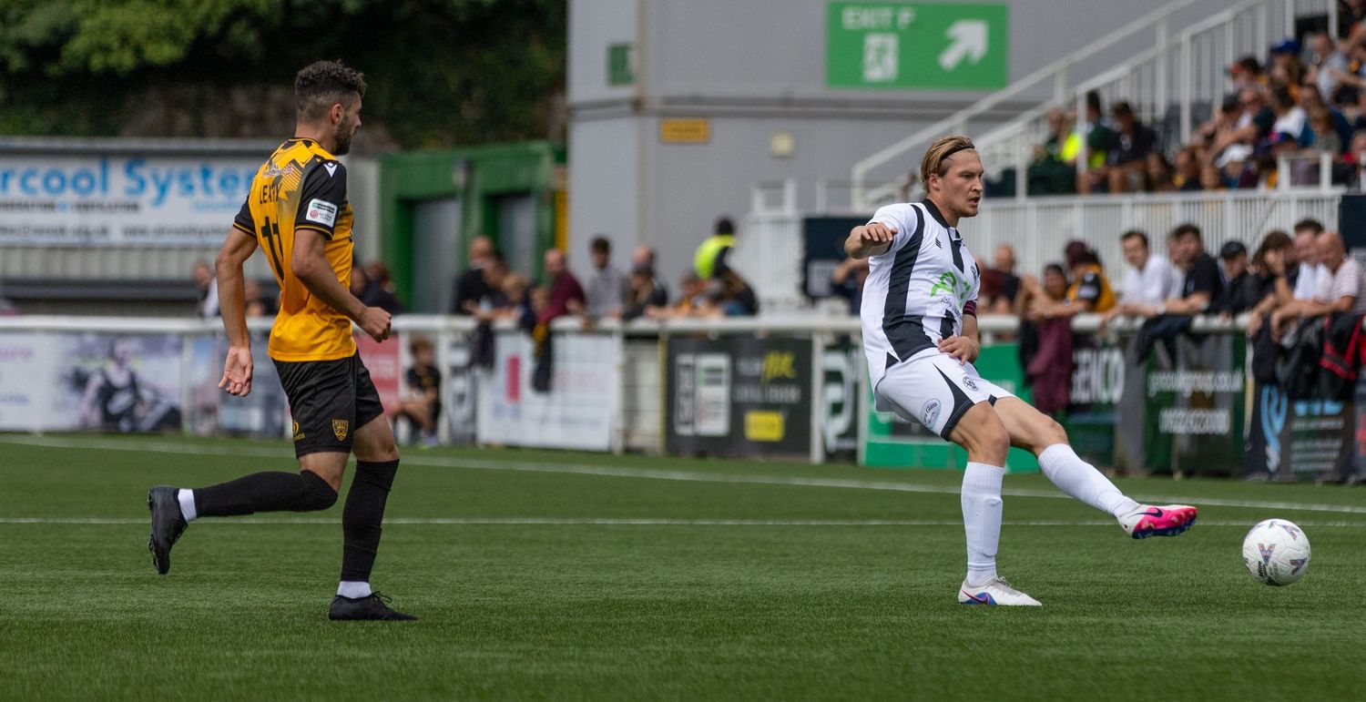 Two soccer players compete on a green pitch, one in white kit striking the ball during a match.
