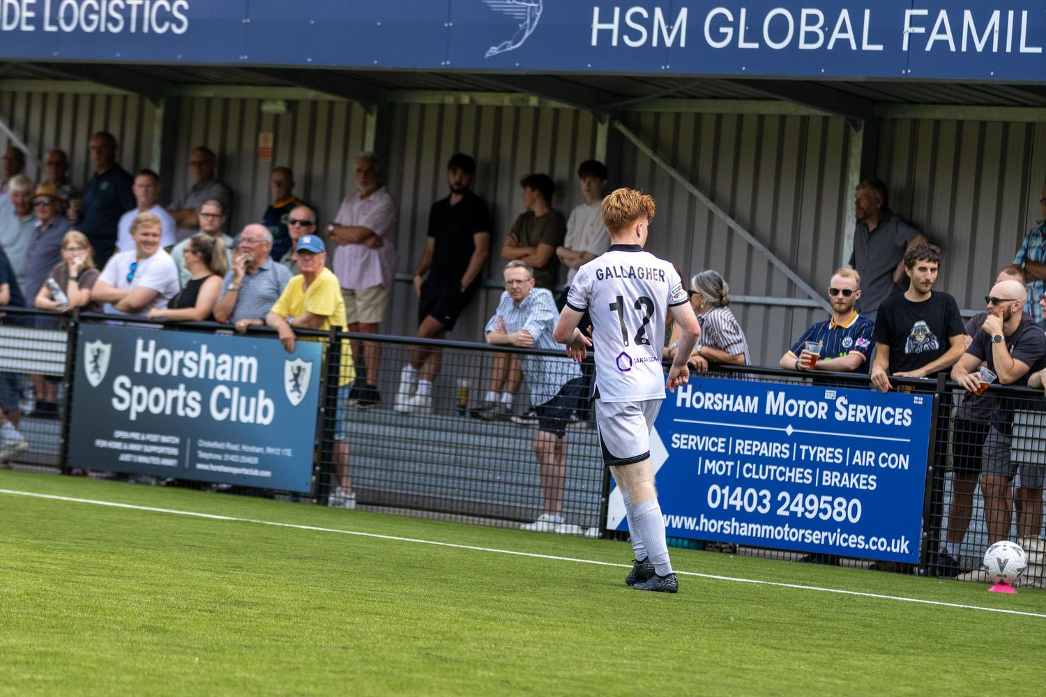 Player number 12 in white kit controls the ball near the sideline at Horsham Sports Club ground.