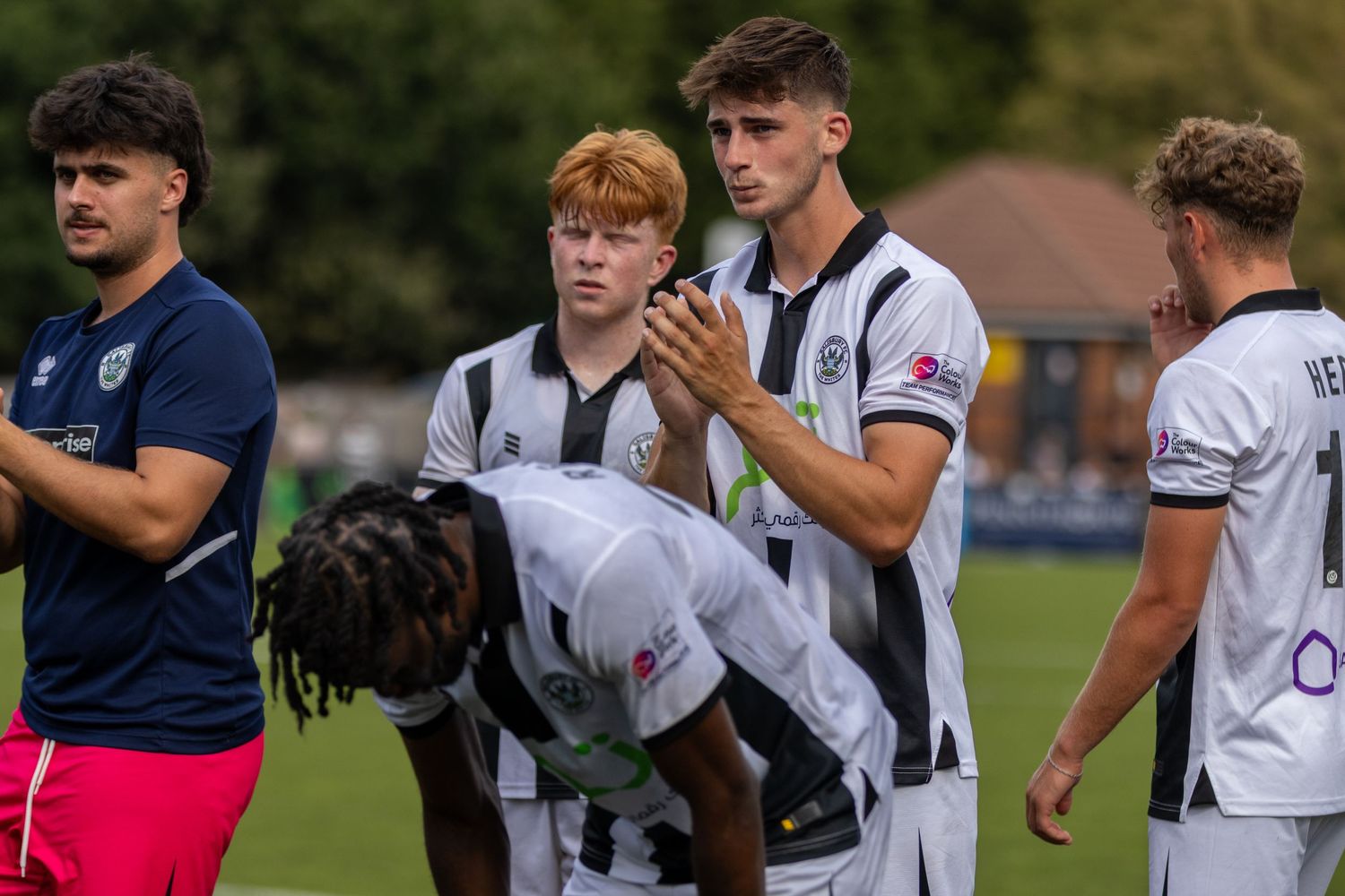 Football players in black and white striped kits applaud fans on the pitch after a summer match.